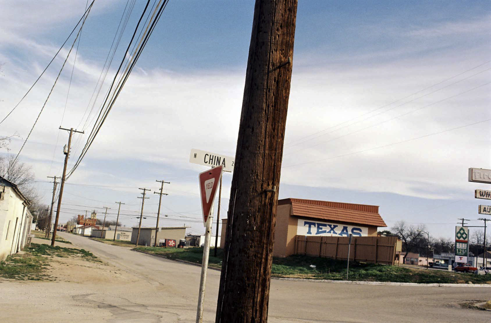 Color photograph of an ex-urban landscape showing an intersection with several low buildings and a row of telephone poles. The nearest building bears a sign with the word "Texas" in blue capital letters; just behind the centermost telephone pole is a street sign reading "China."