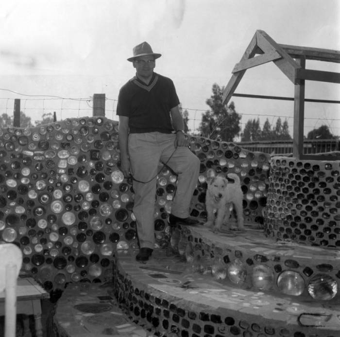 Black-and-white photograph of the same man and dog in fig. 11, standing on curving steps near various structures made of glass bottles.