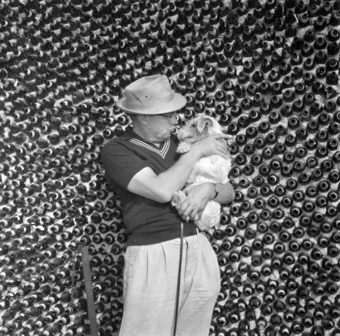 Black-and-white photograph of a man wearing a hat and holding a small dog, standing in front of a wall made from glass bottles.