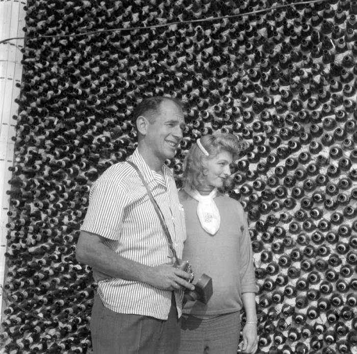 Black-and-white photograph of a man and woman standing in front of a wall made out of glass bottles. The man wears a camera slung across one shoulder.