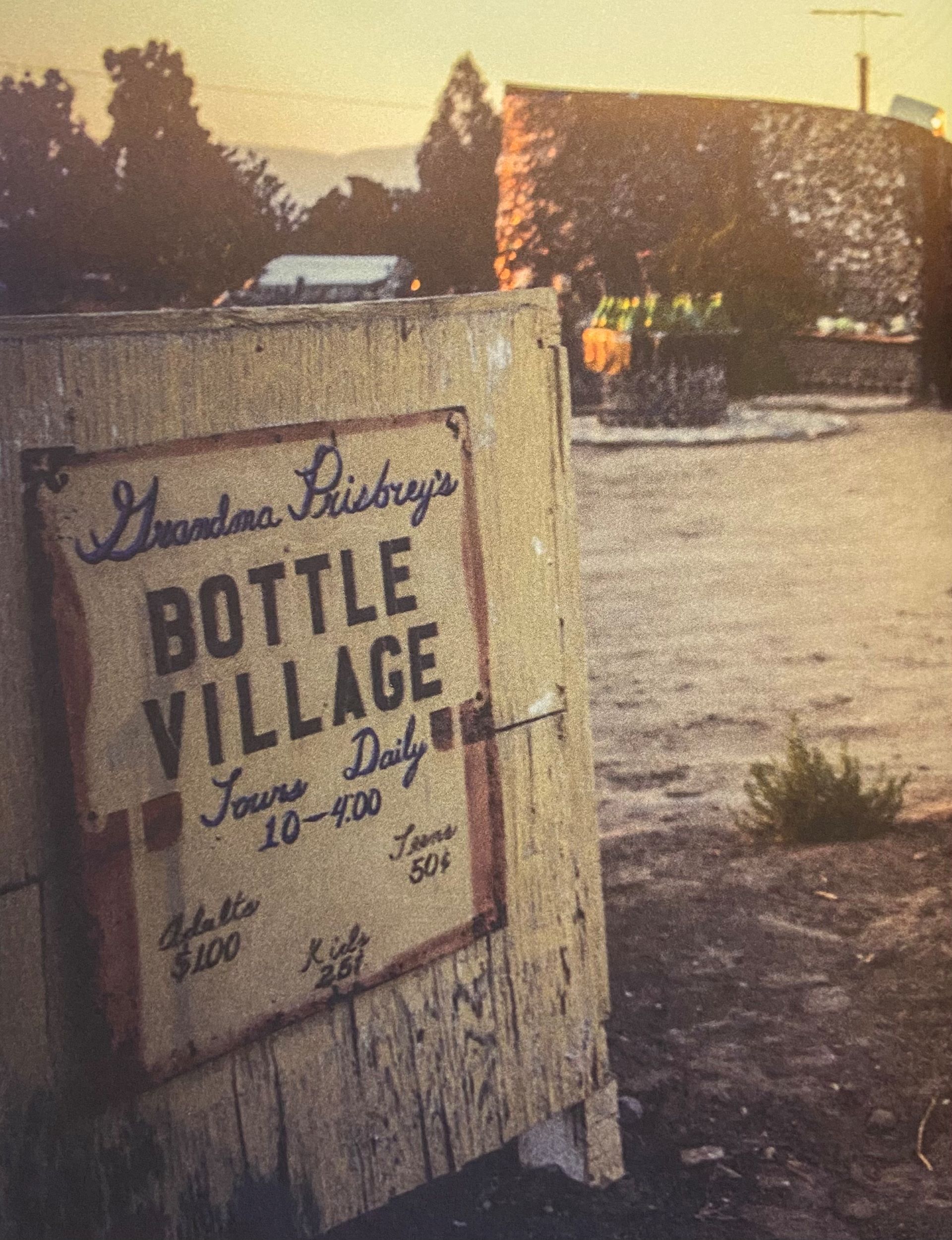 Faded color photograph of a sandwich board-style sign reading "Grandma Prisbrey's / Bottle Village / Tours Daily / 10-4:00," with a view of a structure created from bottles just beyond