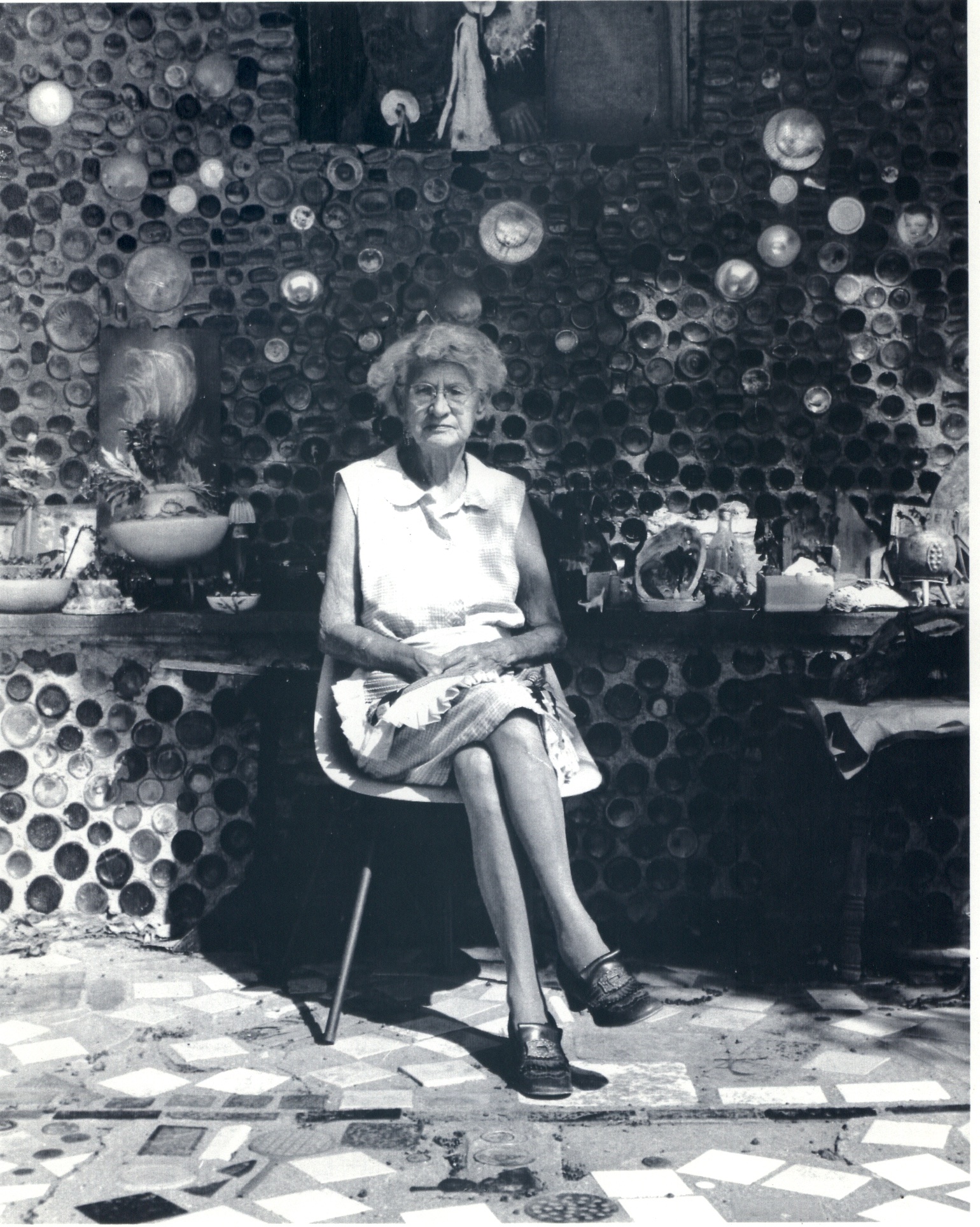 Black-and-white photograph of an elderly white woman wearing a short dress and glasses, seated in front of a structure made from glass bottles