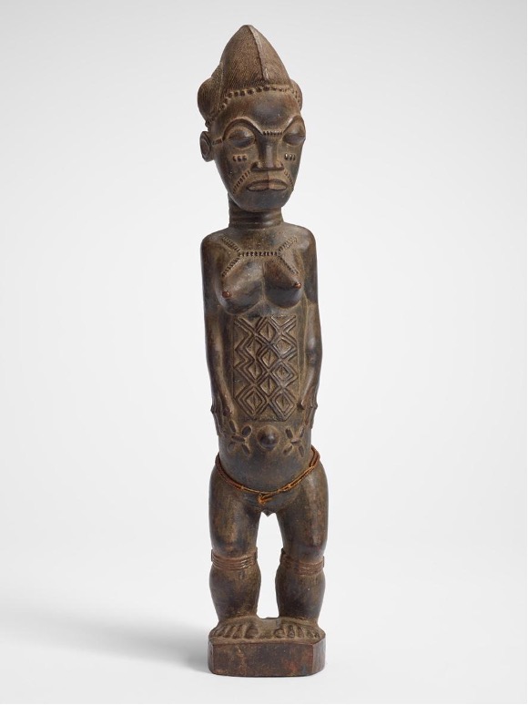 Carved wooden sculpture of a standing human figure with breasts, geometric patterns on the belly, and scarification marks on the face and shoulders.