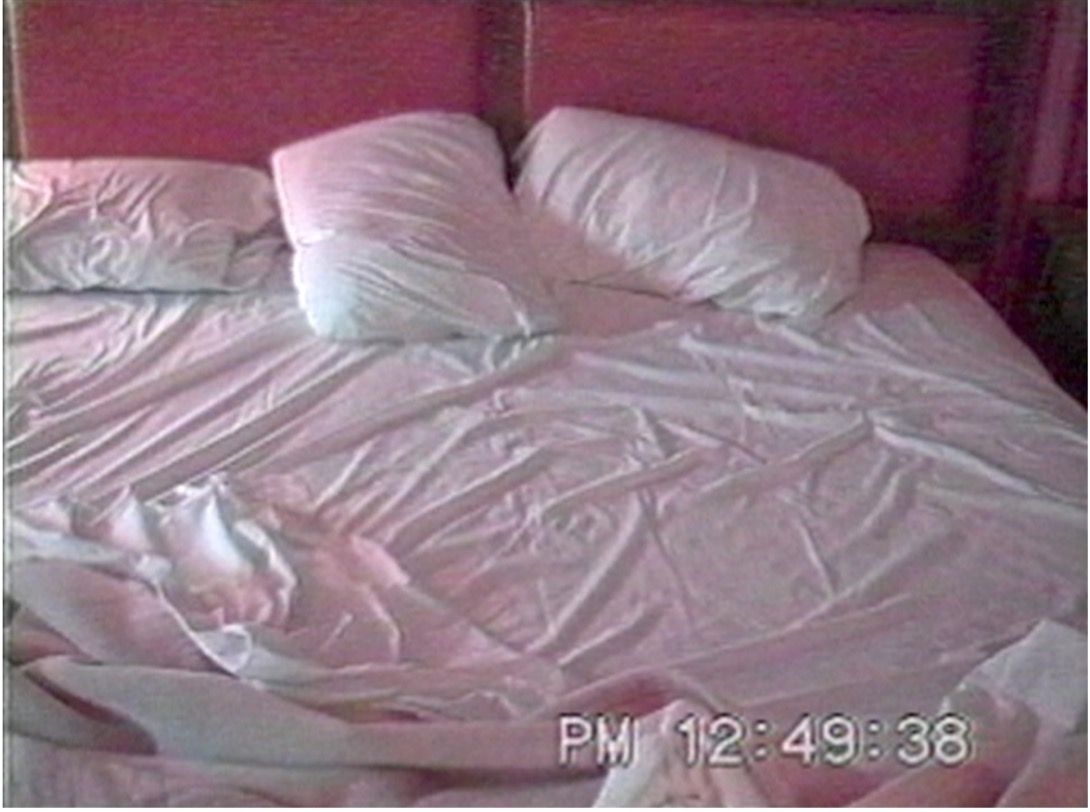 Grainy video still of an unmade bed with rumpled white bedclothes against a deep pink wall. A time stamp reads PM 12:49:38.