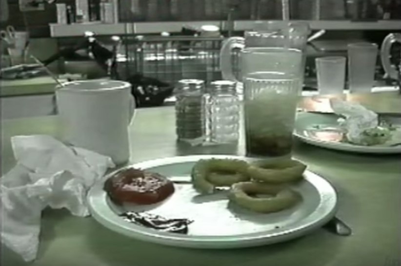 Grainy video still of a table setting at a dinner, showing a partially eaten meal of onion rings and a tomato slice on a white plate, a crumpled napkin, a coffee cup with a spoon in it, a tumbler with ice, and salt and pepper shakers