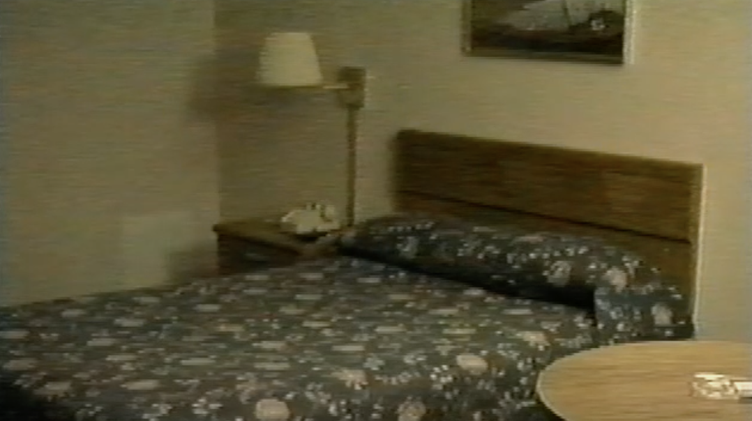 Grainy video still of a hotel room interior, showing a bed with a flowered bedspread and dark wood headboard, a side table with a telephone and a lamp, a framed artwork hanging on the wall, and a round table with an ashtray