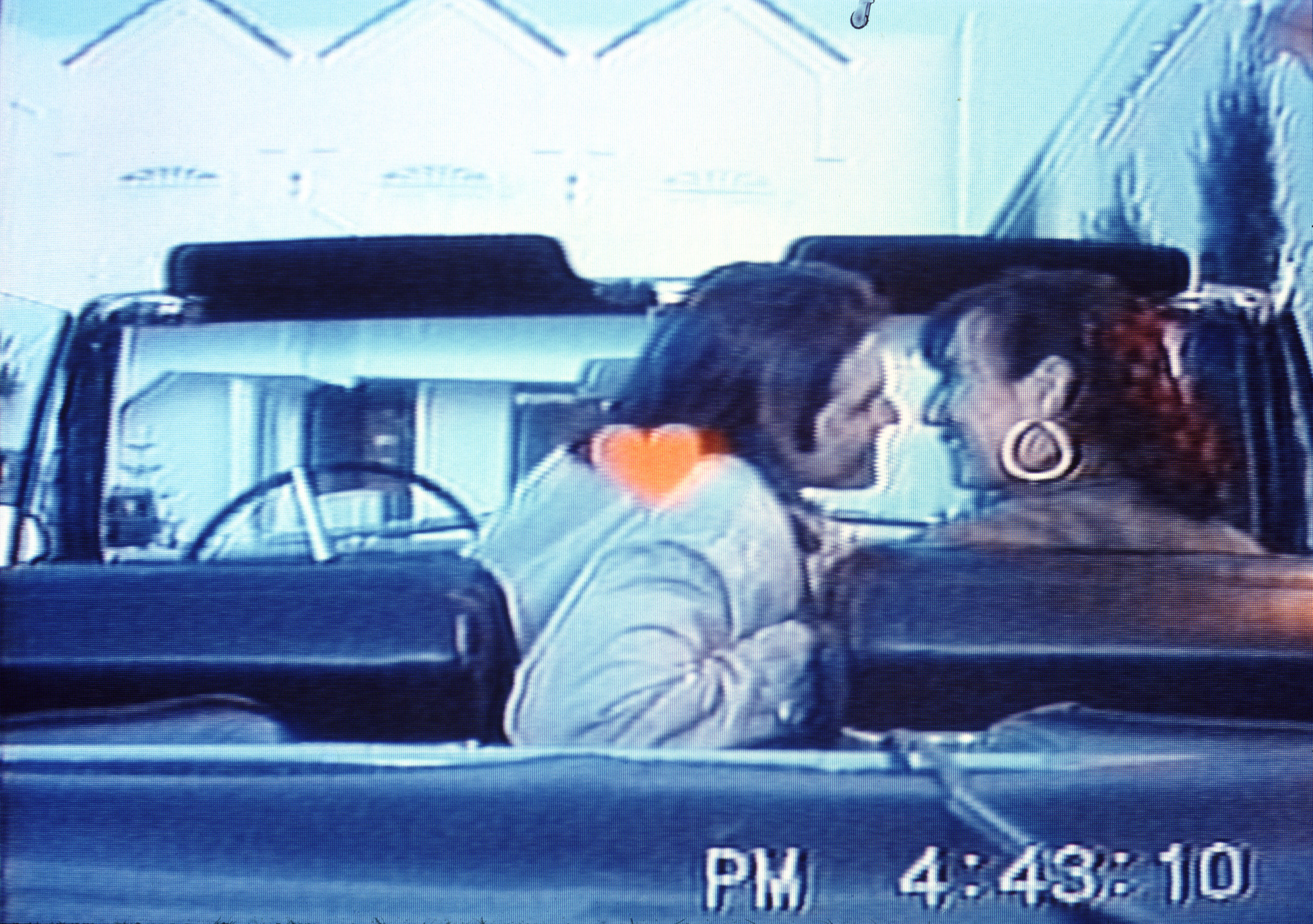 Grainy video still of a man and a woman seen from behind, seated in the front seat of a convertible, with the man leaning over to give the woman a kiss. A small orange heart floats in the center of the image. A time stamp reads PM 4:43:10.