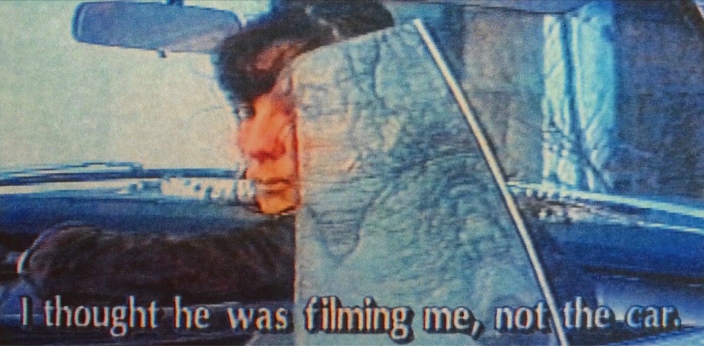 Grainy video still of a woman seated in a car, looking over her shoulder at the viewer. A caption reads, "I thought he was filming me, not the car."