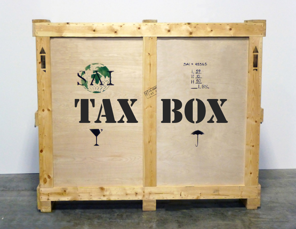 A large crat resting on a gray stone floor. The words "Tax Box," all in capital letters, is stenciled on the crate in black, along with the silhouettes of a martini glass and an umbrella. In the upper left is a green stencil of the globe overlaid with the letters "SAI" in black.