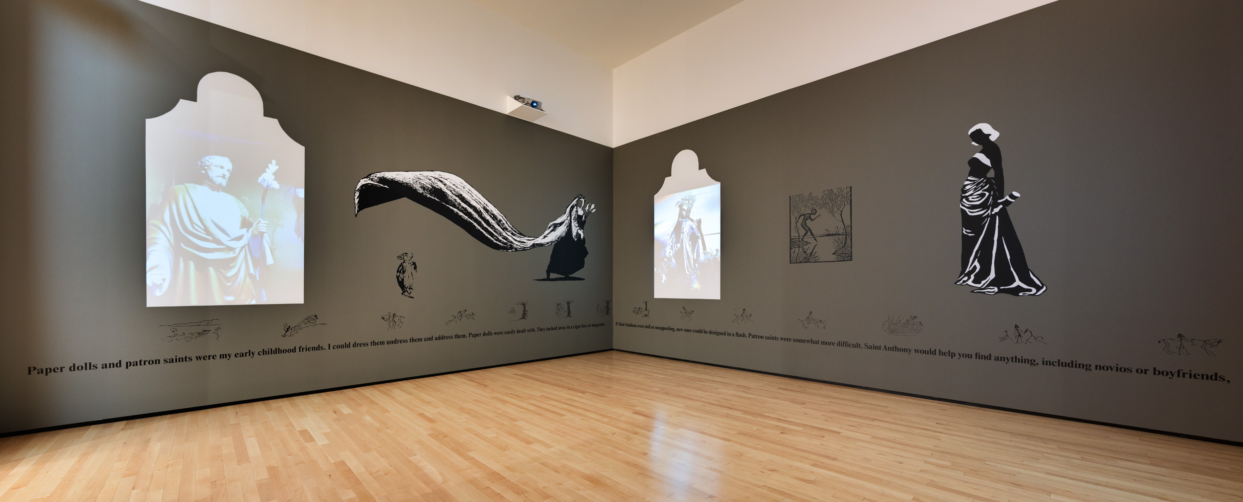 Interior of a museum gallery, where two gray-brown walls meet. On each wall are representations of robed figures combined with text.