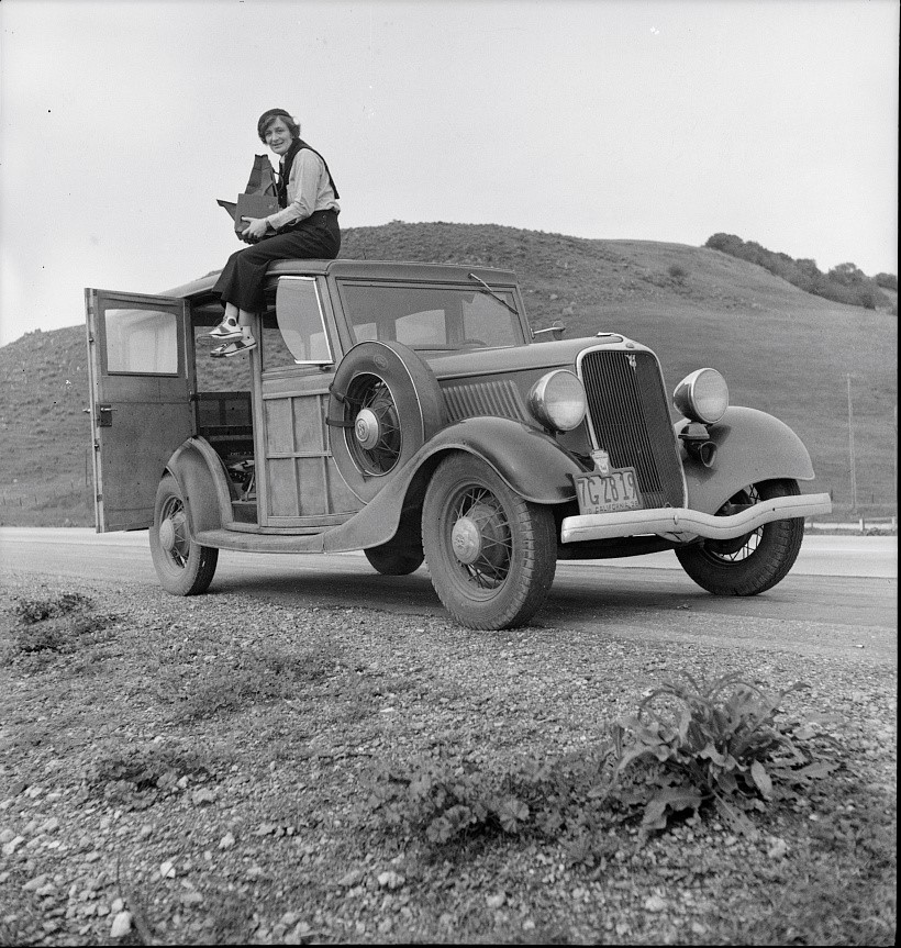 Black-and-white photograph of a woman holding a large camera, seated on the top of a 1930s-era automobile, in front a hill in a rural landscape.