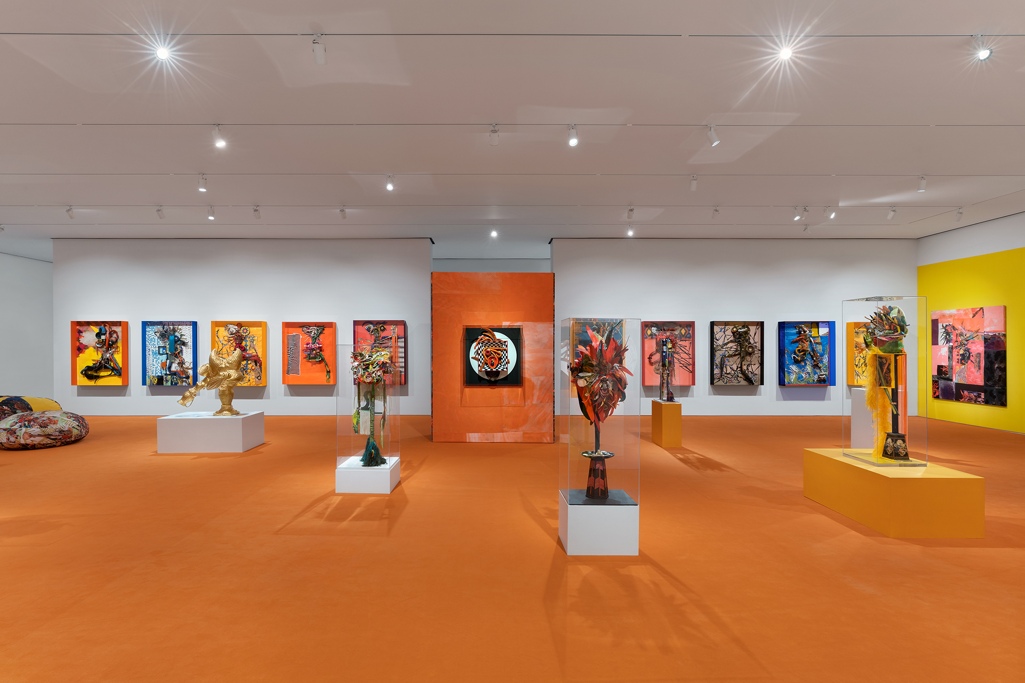 Interior of a museum gallery, showing eleven colorful framed works hung on the walls and five freestanding sculptural pieces on the orange-colored floor.