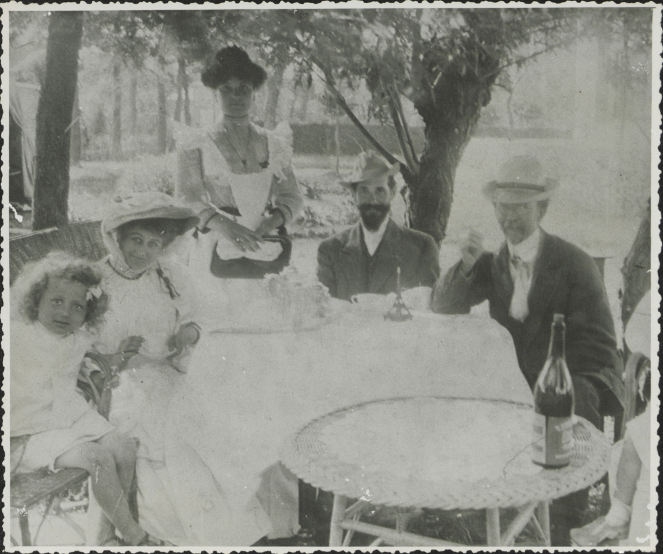 Black-and-white photograph of five people seated outdoors before a table covered in a white cloth: two women, two men, and one young girl. 