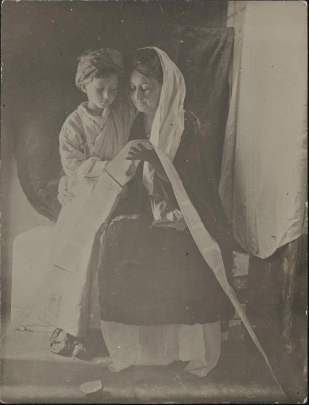 Black-and-white photograph of a seated woman with a white cloth draped over her head, embraced by a young child wearing a white robe. Together they are reading something on a long scroll that drapes over the woman's lap.