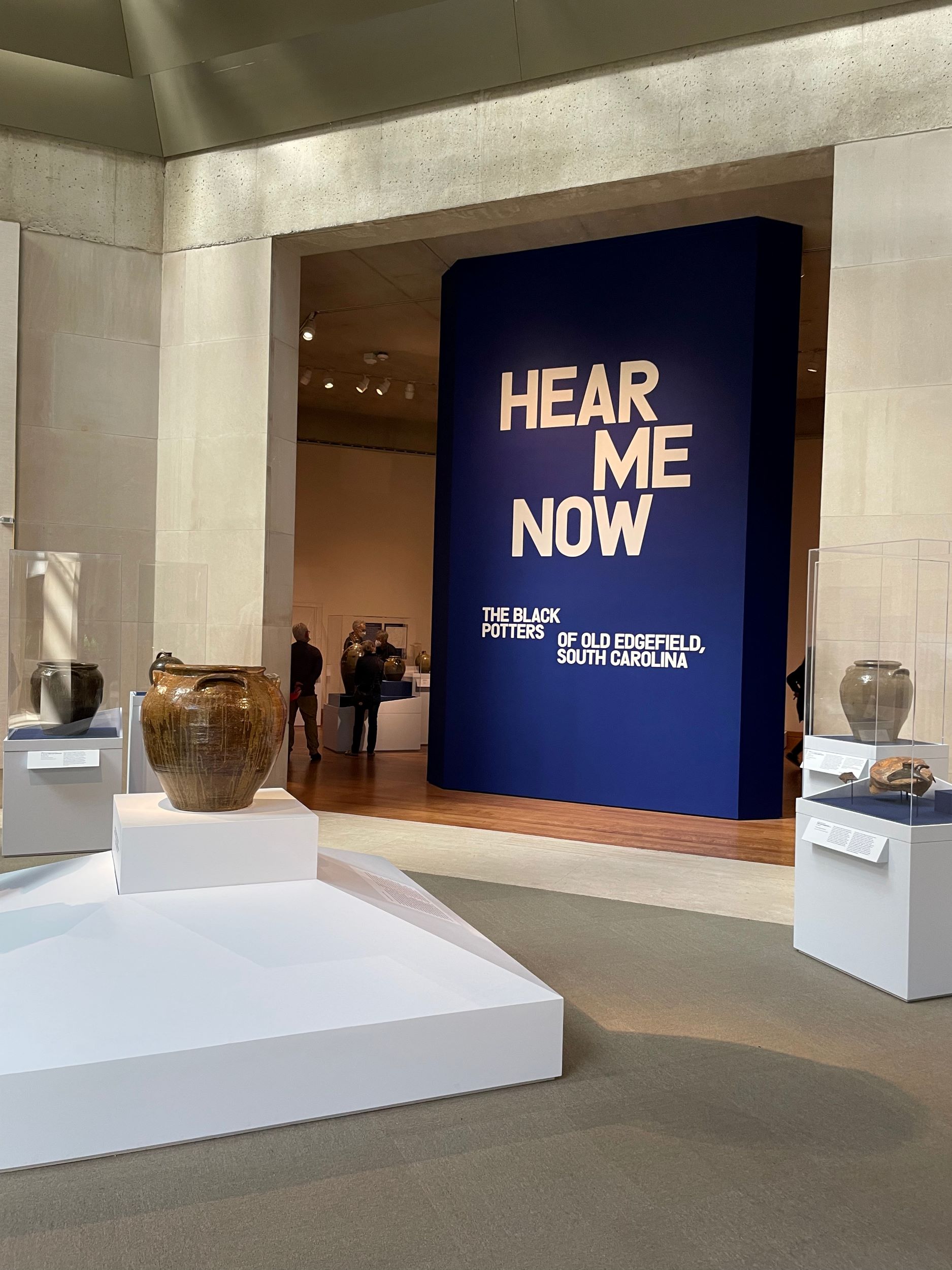 Entrance to a museum exhibition, with a freestanding wall that reads, in white, "Hear Me Now / The Black Potters / of old Edgefield, / South Carolina." In front of it are several pedestals with ceramic vessels resting on them.