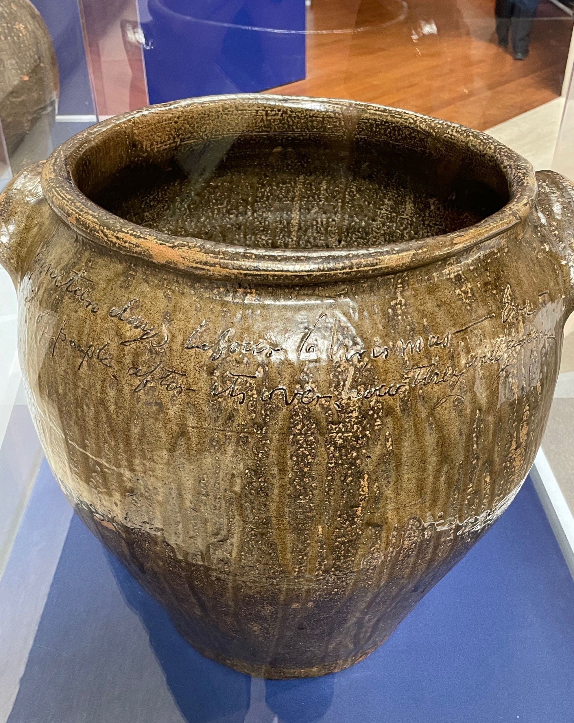 Large ceramic jug with a shiny, uneven brown glaze. A largely illegible script inscription runs under the rim.
