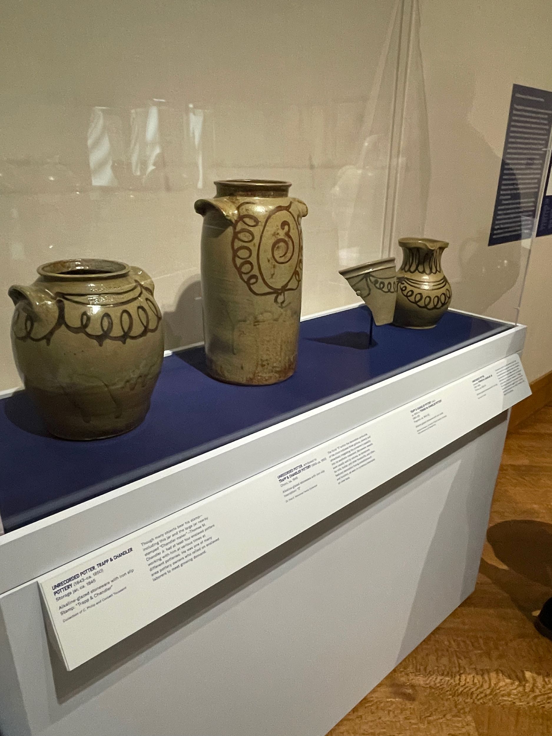 A glass-enclosed museum case featuring three ceramic vessels, with curlicued slip decorations, resting on a deep blue cloth. The case also includes a vessel fragment. 