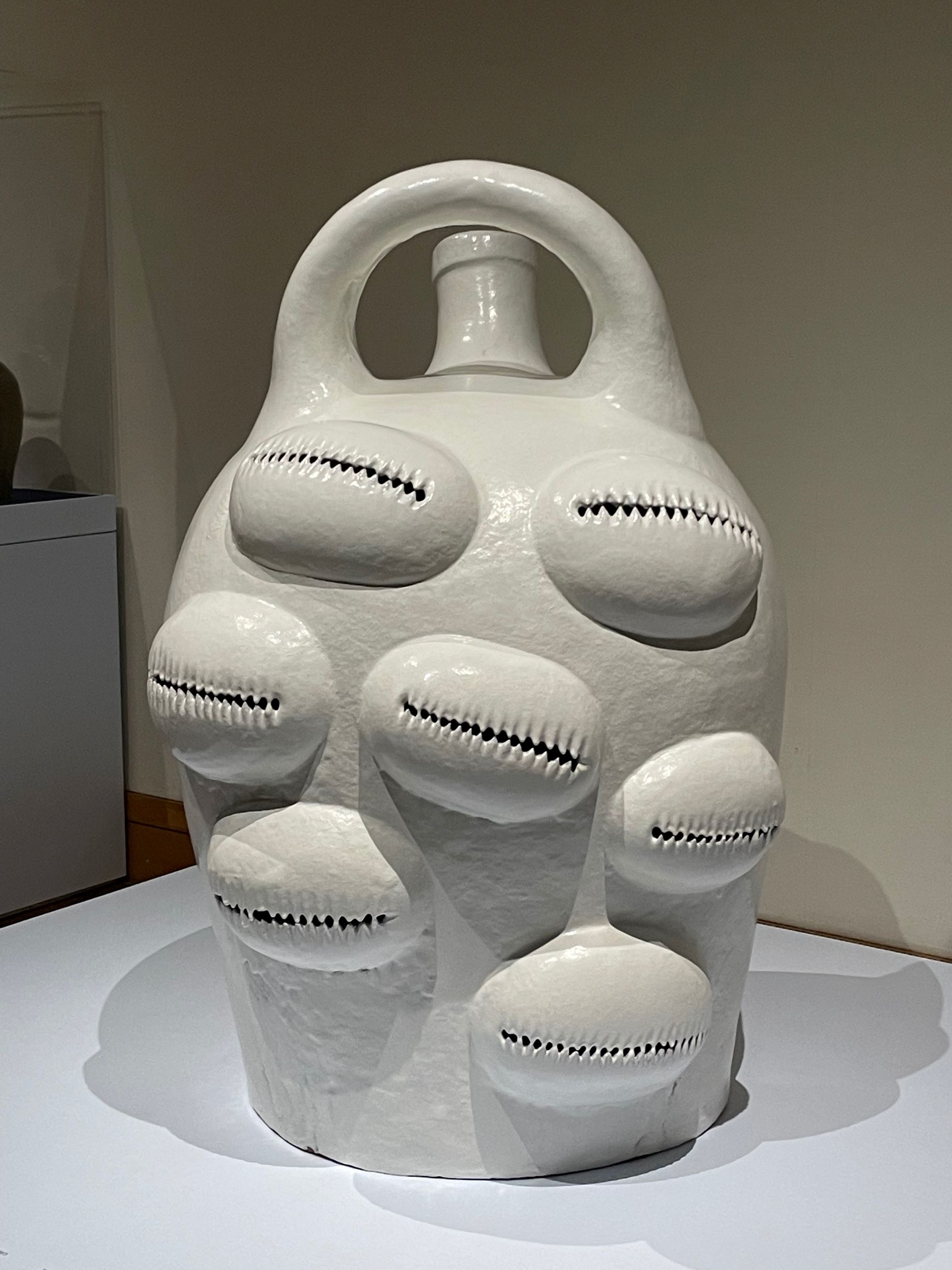 A white-glazed ceramic vessel with a handle and spout. Seven large shapes that resemble cowrie shells are embedded on the form.