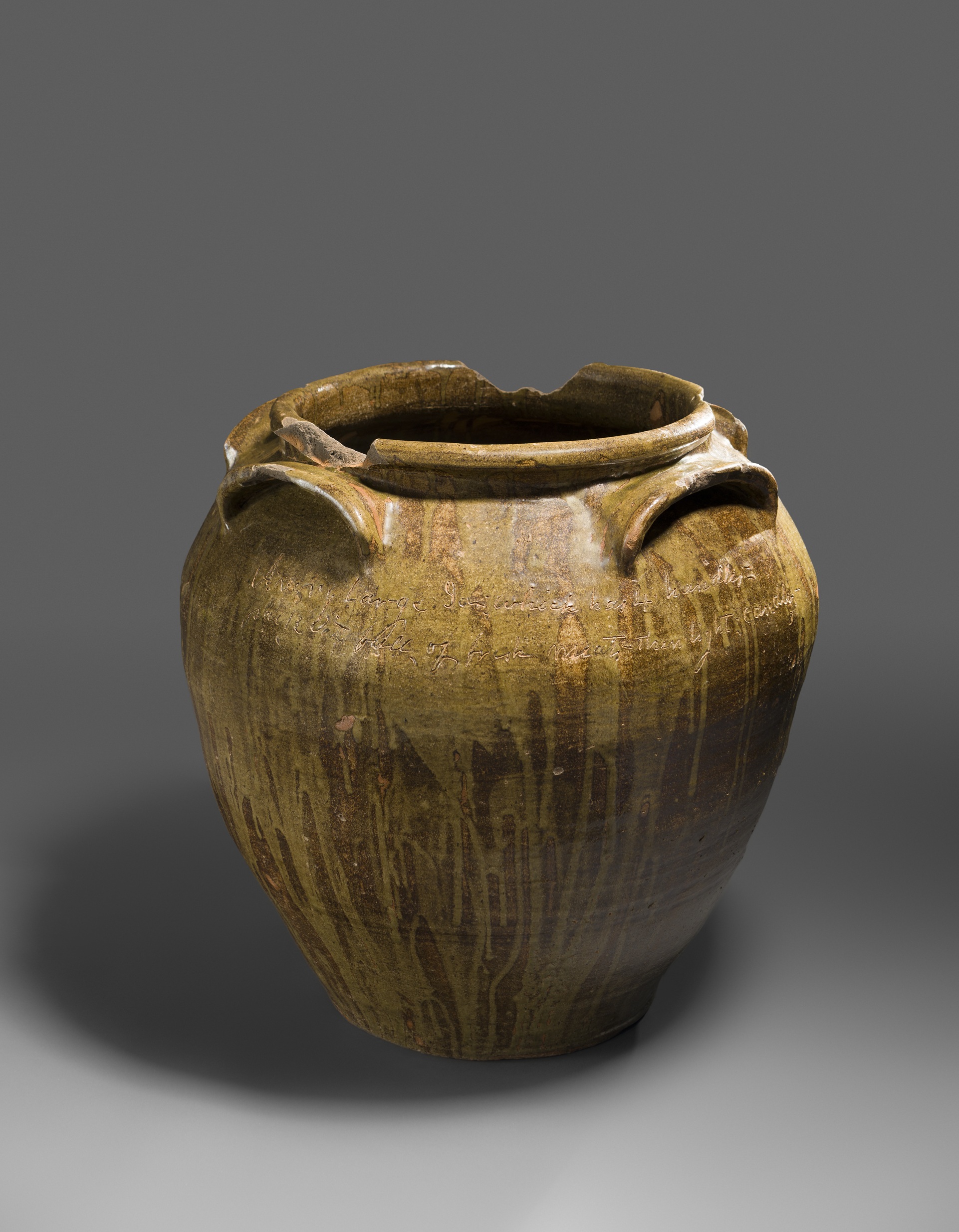 Ceramic vessel with wide mouth and four spoon-shaped handles. The vessel is covered in a variegated brown glaze. The rim has several chips missing from it.