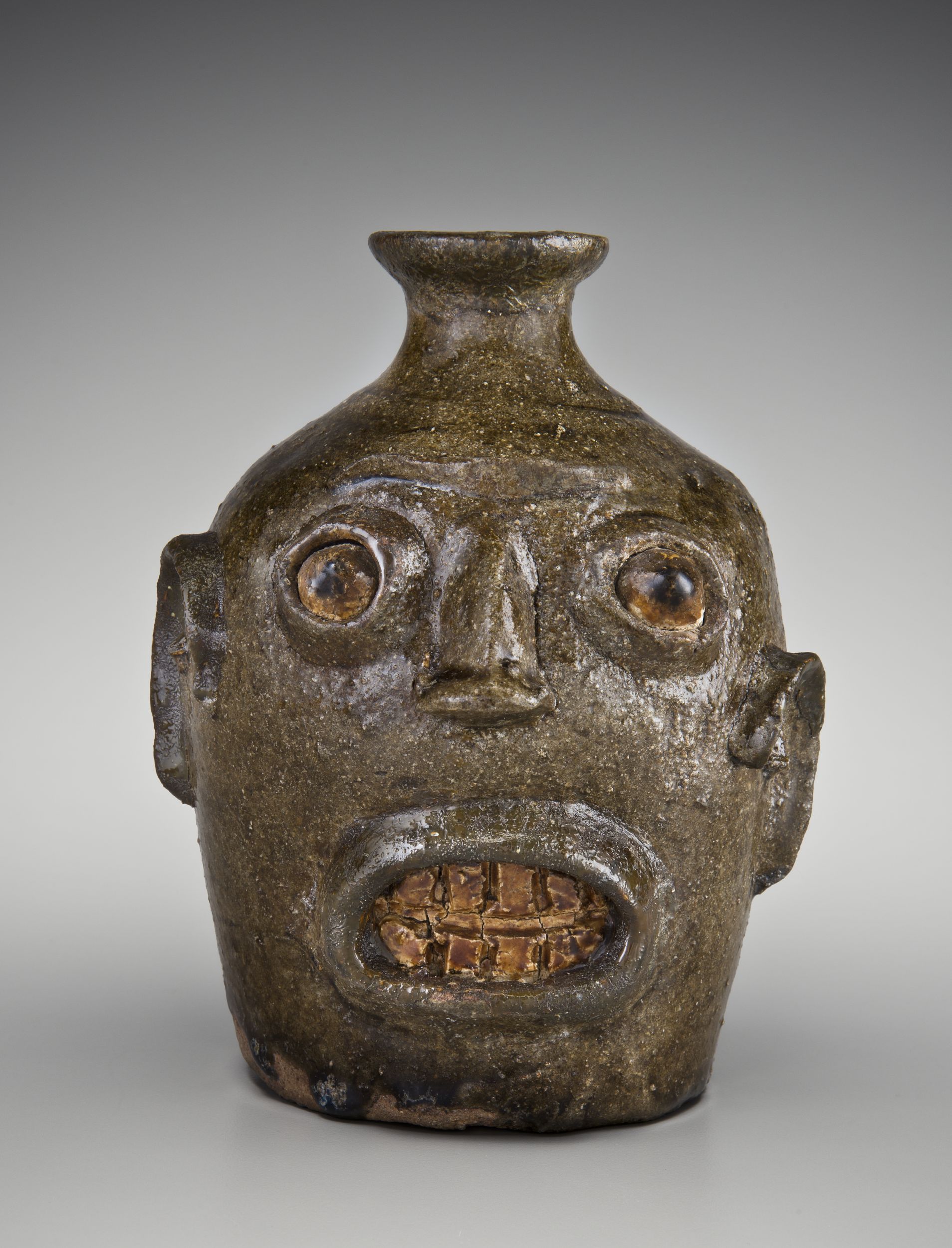 Ceramic jug in the form of a human face, with wide-open eyes and an open mouth with visible teeth. The vessel is finished in a rough, dark glaze.
