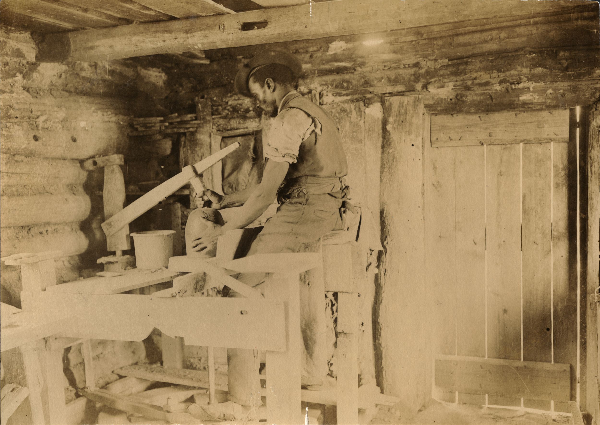 Sepia-toned photograph of a Black man inside a log cabin, wearing shirtsleeves and a vest, seated at a wooden pottery wheel with a ceramic vessel in front of him.