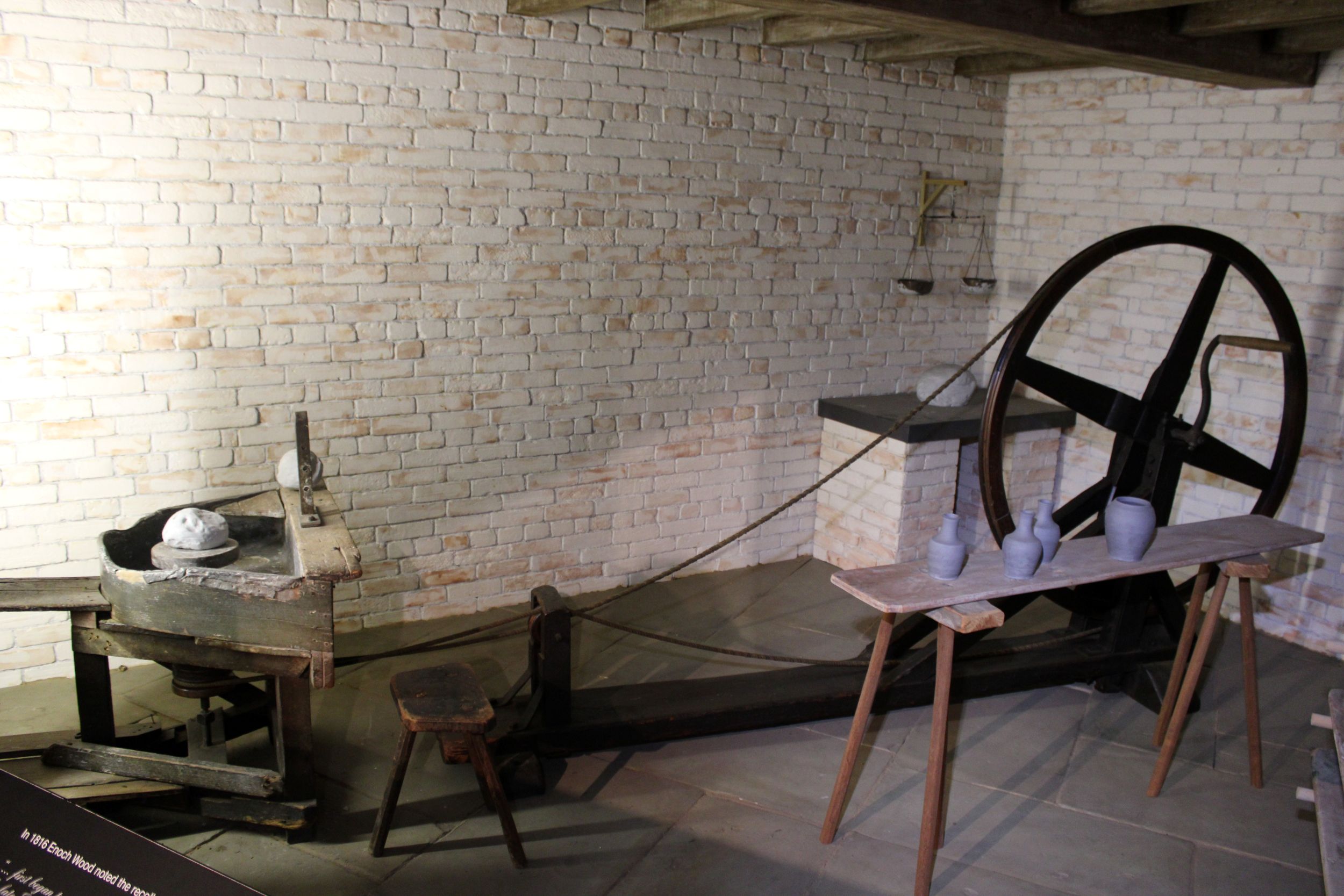 Interior of a room faced with white brick, with a large potter's wheel and a bench with several ceramic vessels on the left.