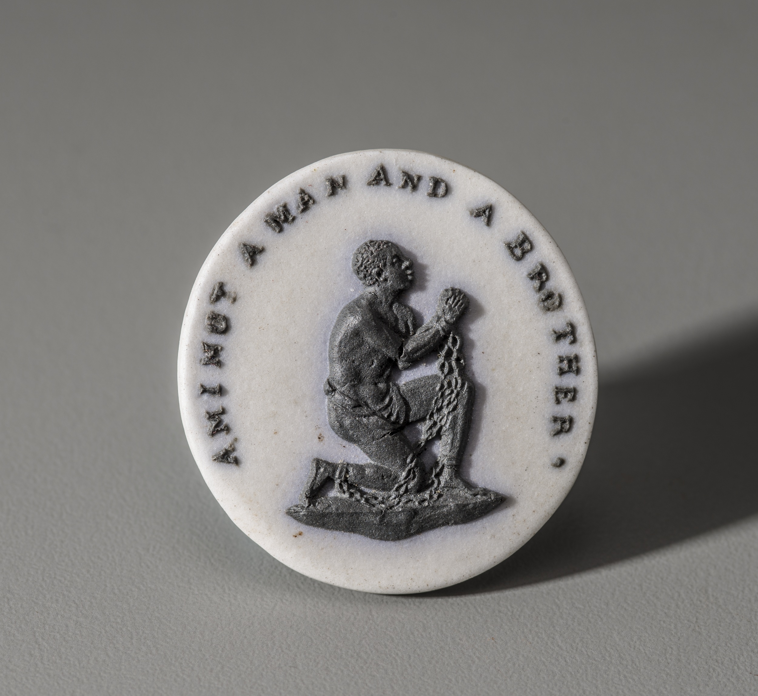 Medallion with a white ceramic background, against with is a kneeling figure, rendered in black, wearing chains and clasping his hands in prayer. Arching over his head are the words "Am I not a man and a brother"