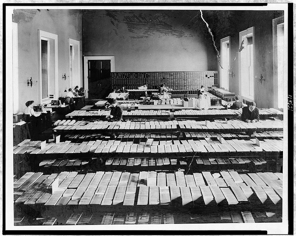 Black-and-white photograph of a large room filled with tables covered with an open filing system, lit by windows to the right. Several white people are working with the files, and an old-fashioned library card catalogue stands in the back of the room.