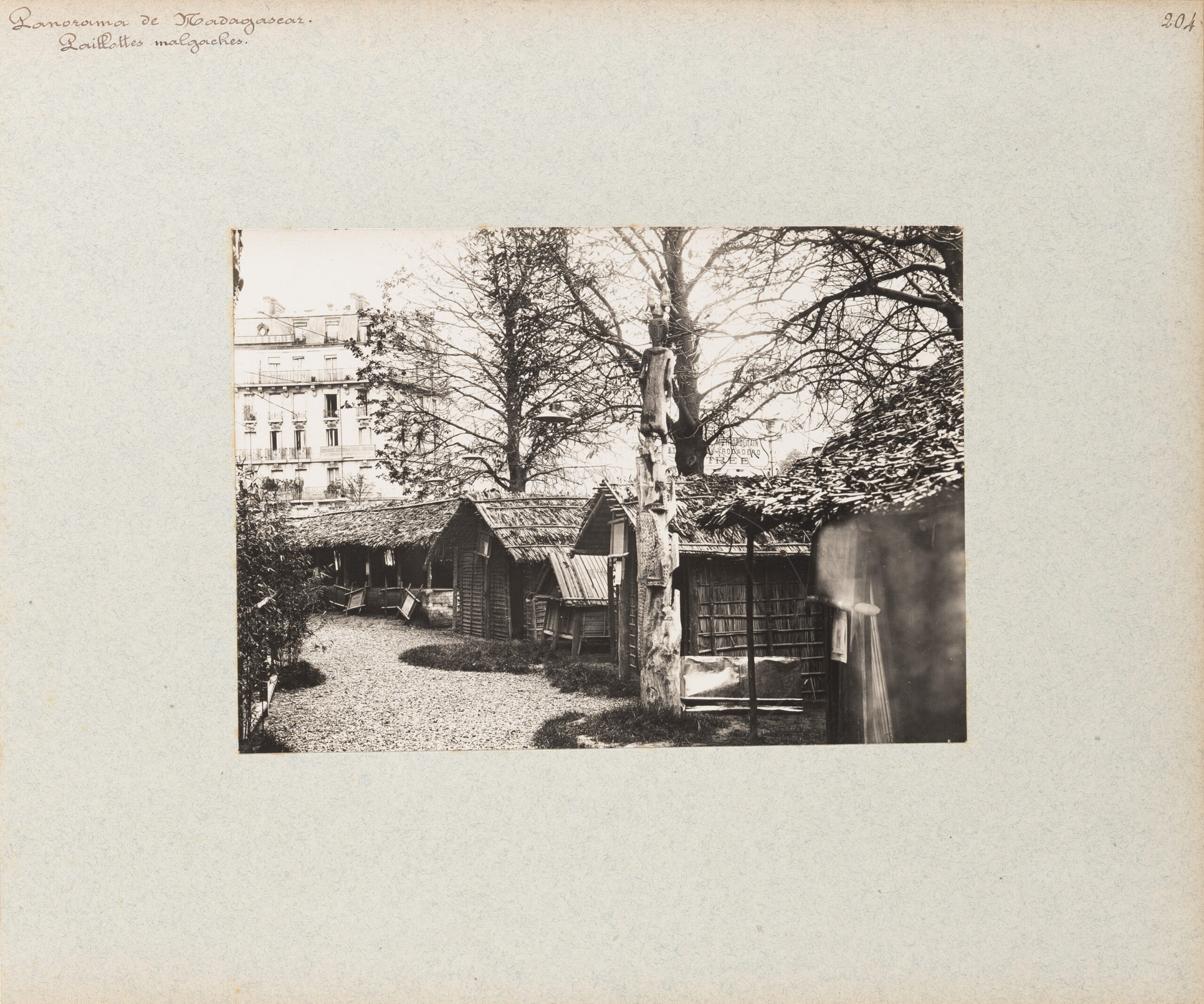 Black-and-white photograph, mounted on matboard, depicting a series of low, shed-like buildings with thatch-like roofs, with trees beyond. In the distance to the left is a larger multistory building. The cursive inscription on the upper left of matboard reads "Panorama de Madagascar. / Paillottes malgaches"