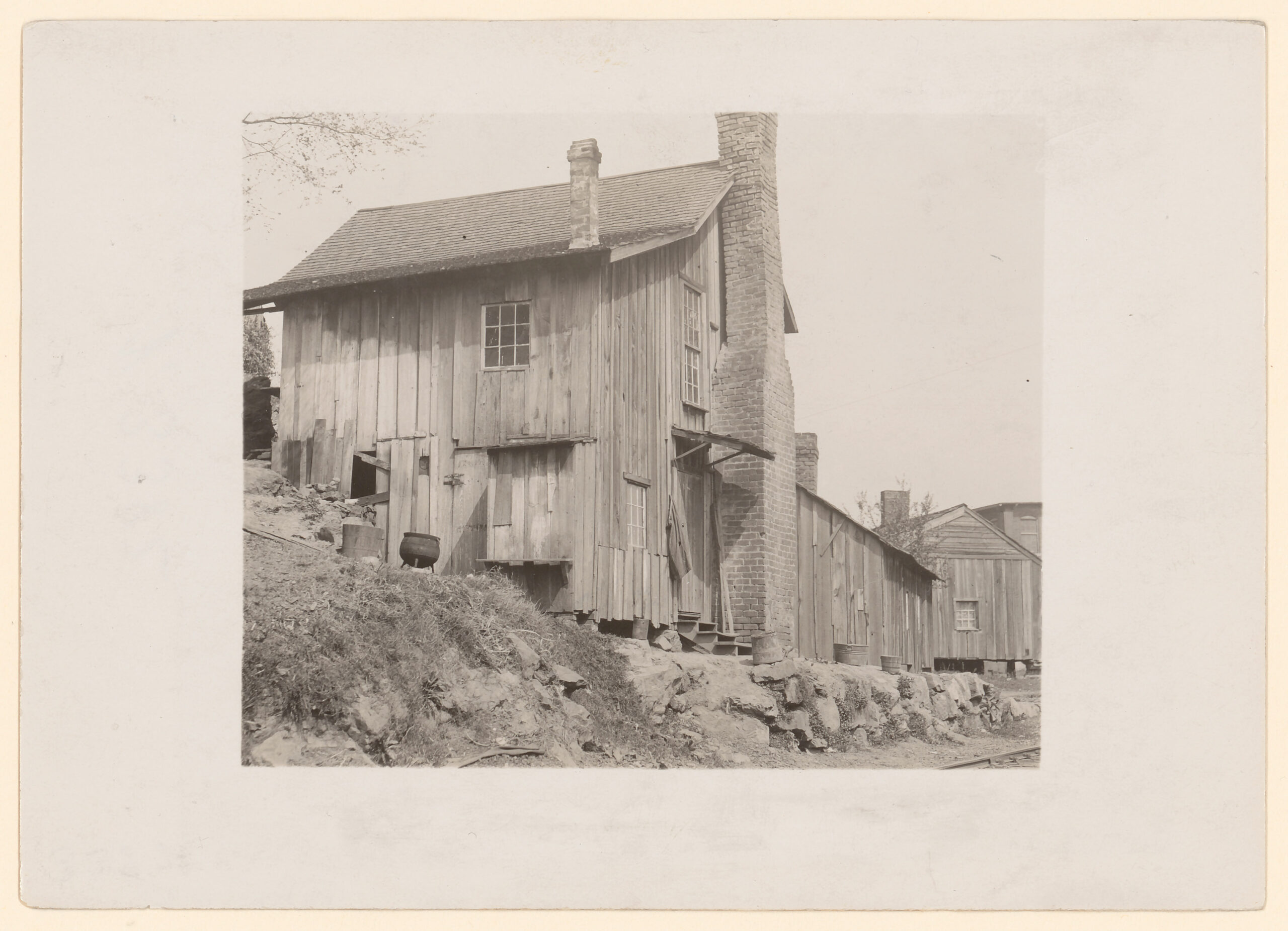 Black-and-white photograph, mounted on matboard, of a shack with vertical clapboards, two visible windows, a shingled roof, and a brick chimney, clinging to the side of a scrubby hill.