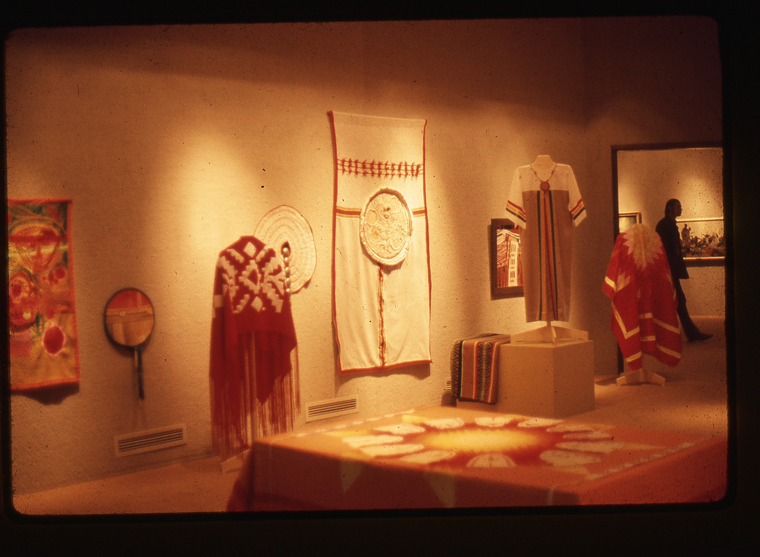 Aged color photographic image of a museum exhibition featuring Native American textiles, including several wall hangings, three freestanding garments, and one textile spread over a low, wide plinth. Far to the right is a doorway through which can be seen the silhouette of a museum visitors, with traditional paintings behind them.