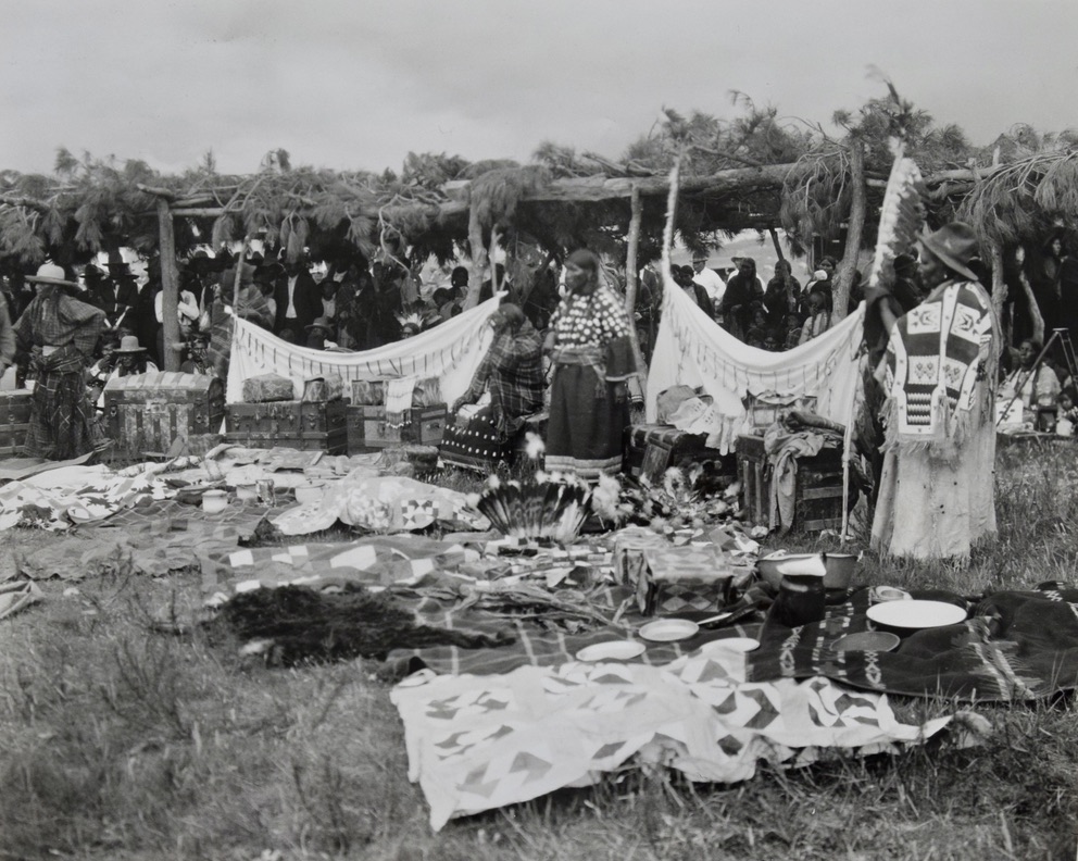 Black-and-white photograph showing several Native American men and women standing behind textiles and other objects spread out over the grass.