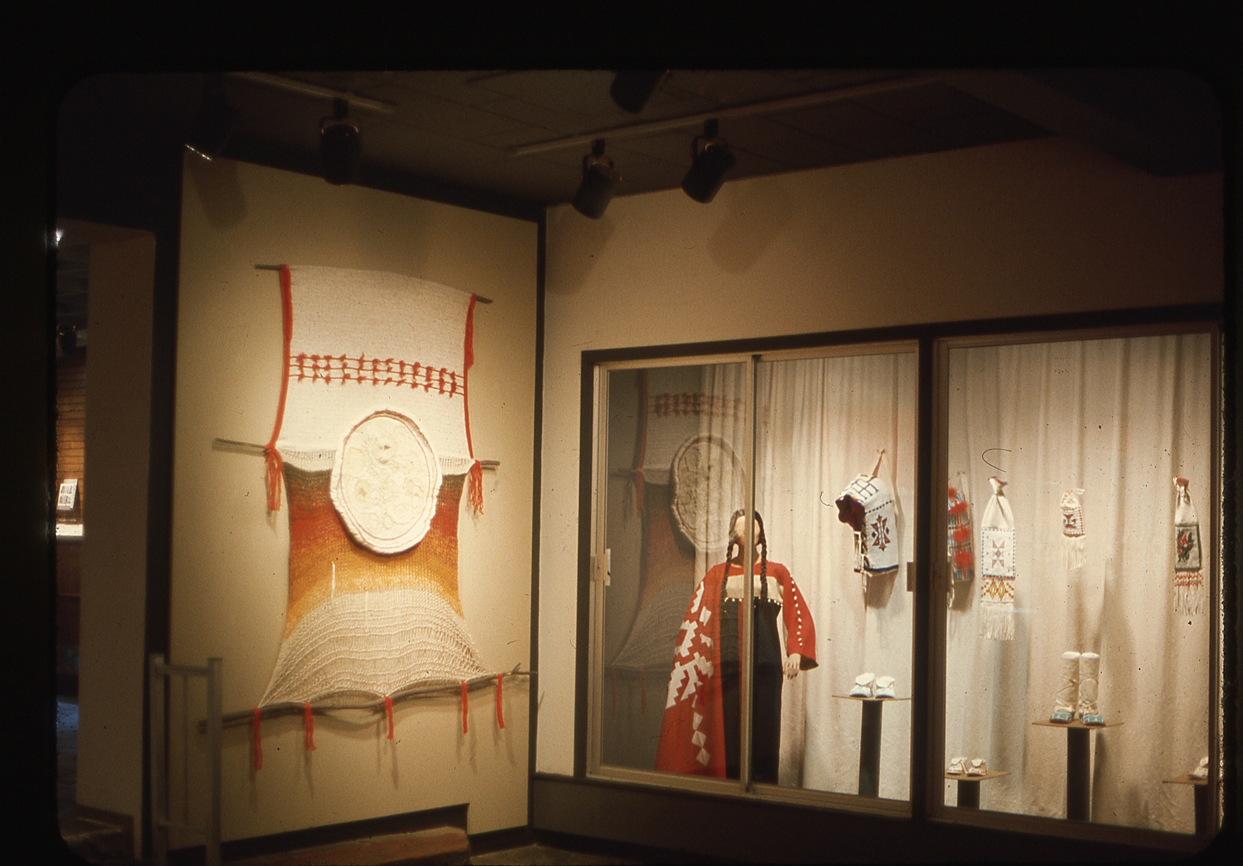 Aged color photograph of a museum exhibition of native American arts. on the left is a large woven wall hanging; on the right is a glass wall, behind which are examples of cloaks, headgear, and footwear.