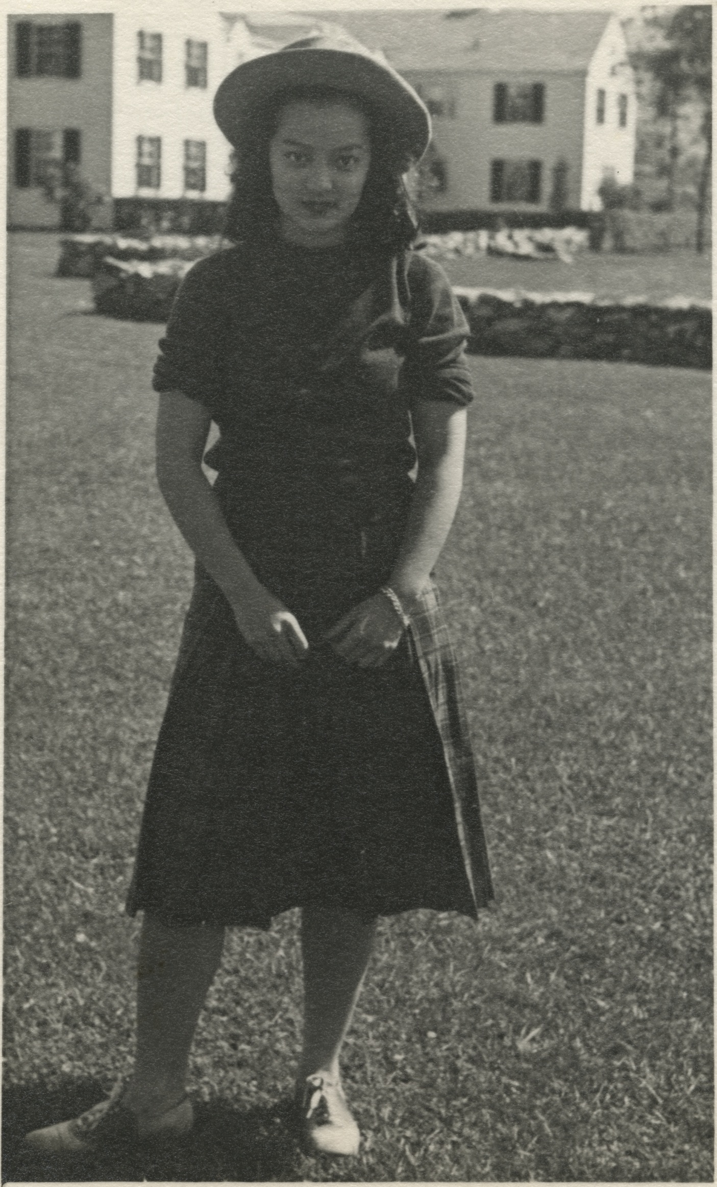 Black-and-white photograph of a young woman wearing a plaid skirt, a wide-brimmed hat, a short-sleeved sweater, and saddle shoes, standing on a lawn in front of a colonial-style house with a stone fence.
