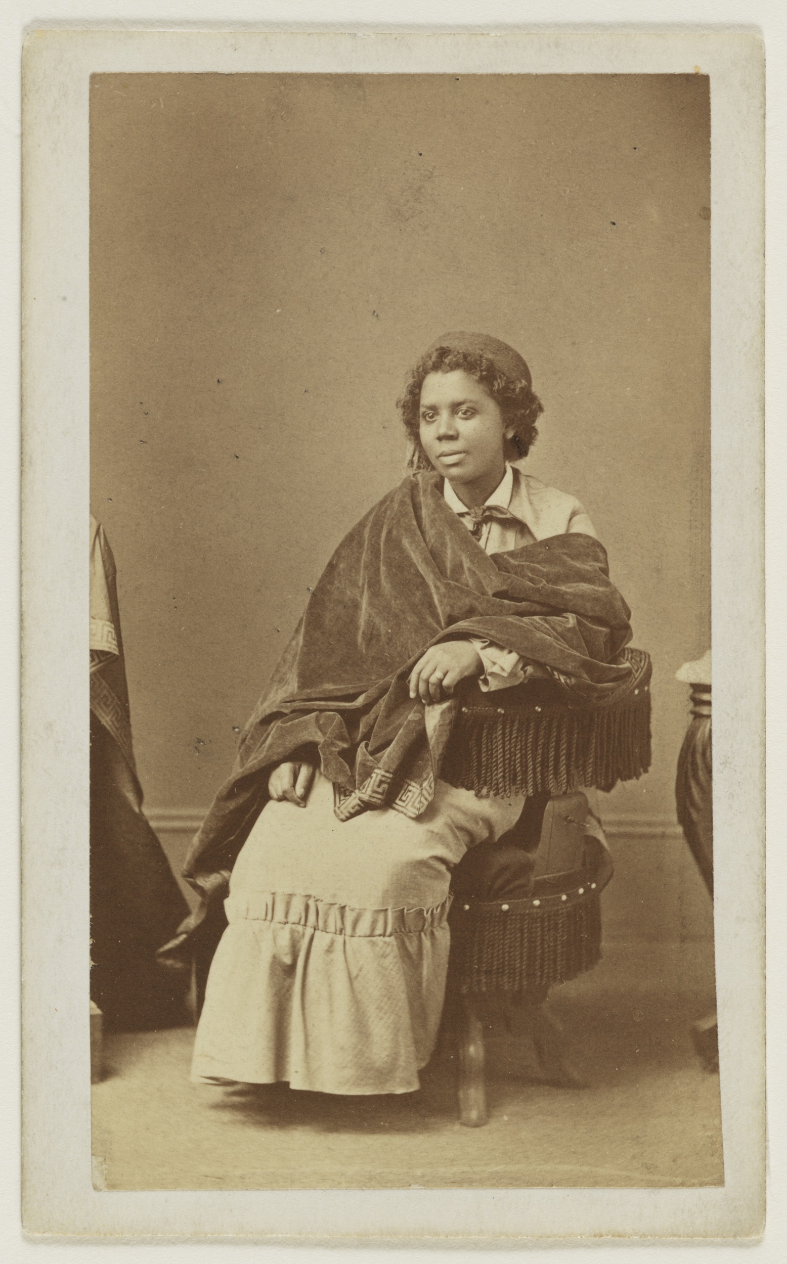 Sepia-toned photograph of a Black woman seated on a chair, wearing a pillbox-style cap, a long dress, and a velvet shawl wrapped around her upper body.