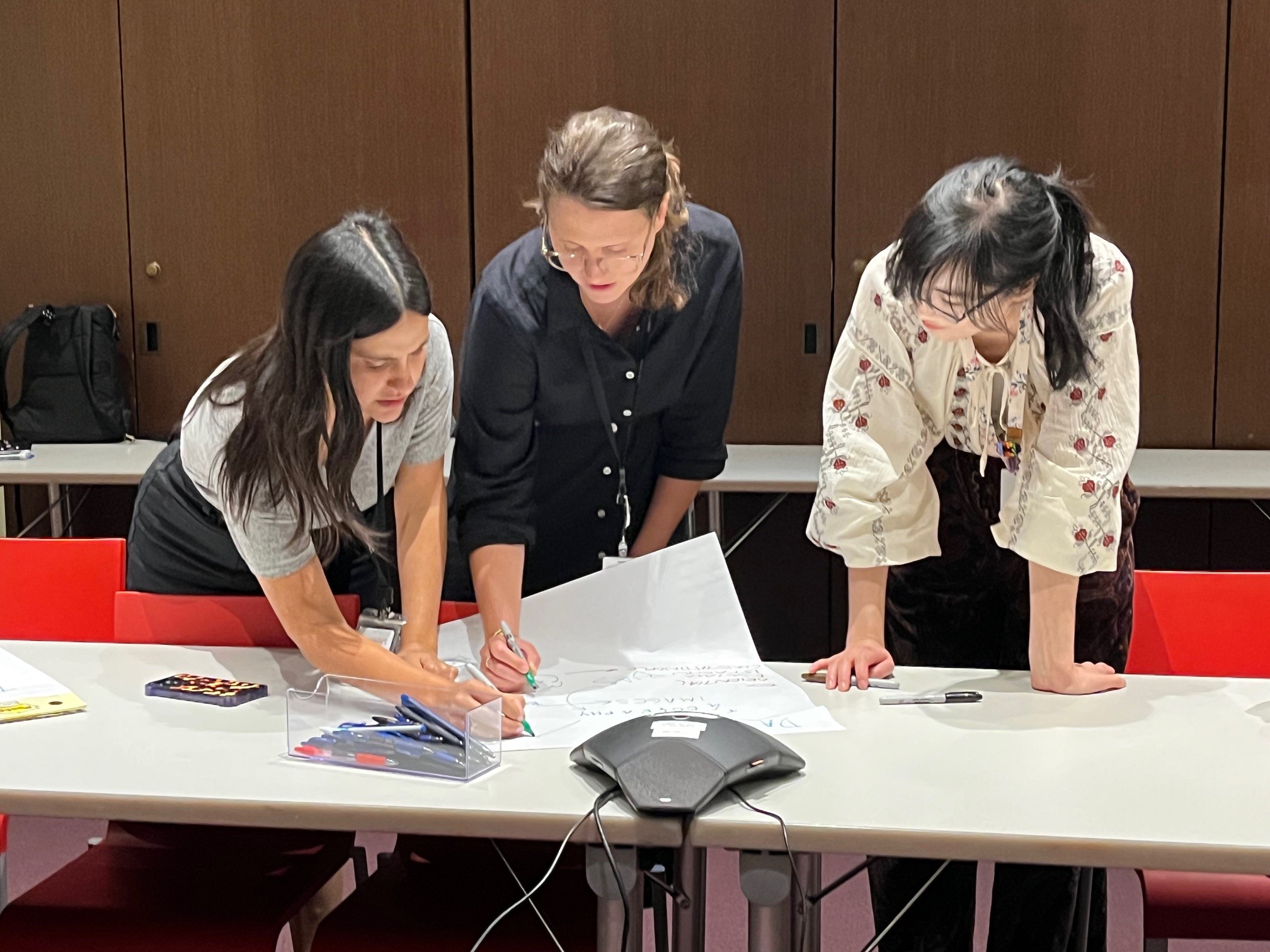 Three women in a conference room leaning over a table, marking a large sheet of paper together.
