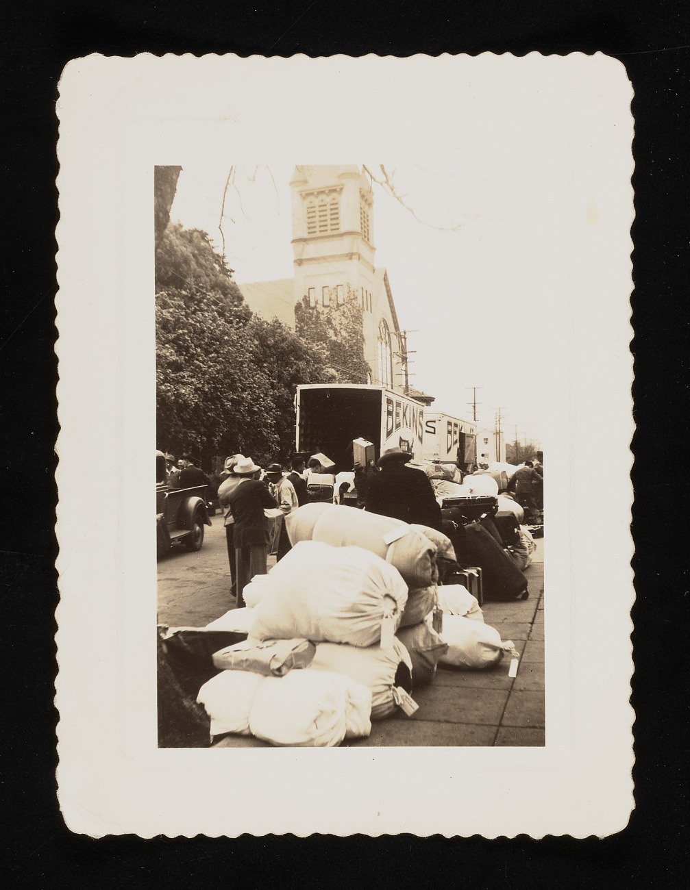 Black-and-white photograph with deckled edges, showing large white sacks lying on a sidewalk in front of two open trucks labeled "Bekins." Dense bushes and the facade of a church are visible in the background.