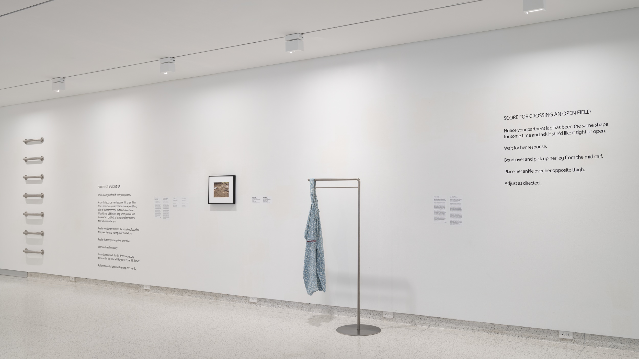 Interior of a museum gallery with white walls. To the left, seven grab bars are attached to the wall in a vertical line; in the center is a small framed work, and on the left is a blue garment hung on a bent metal stand. There is illegible text on the walls.
