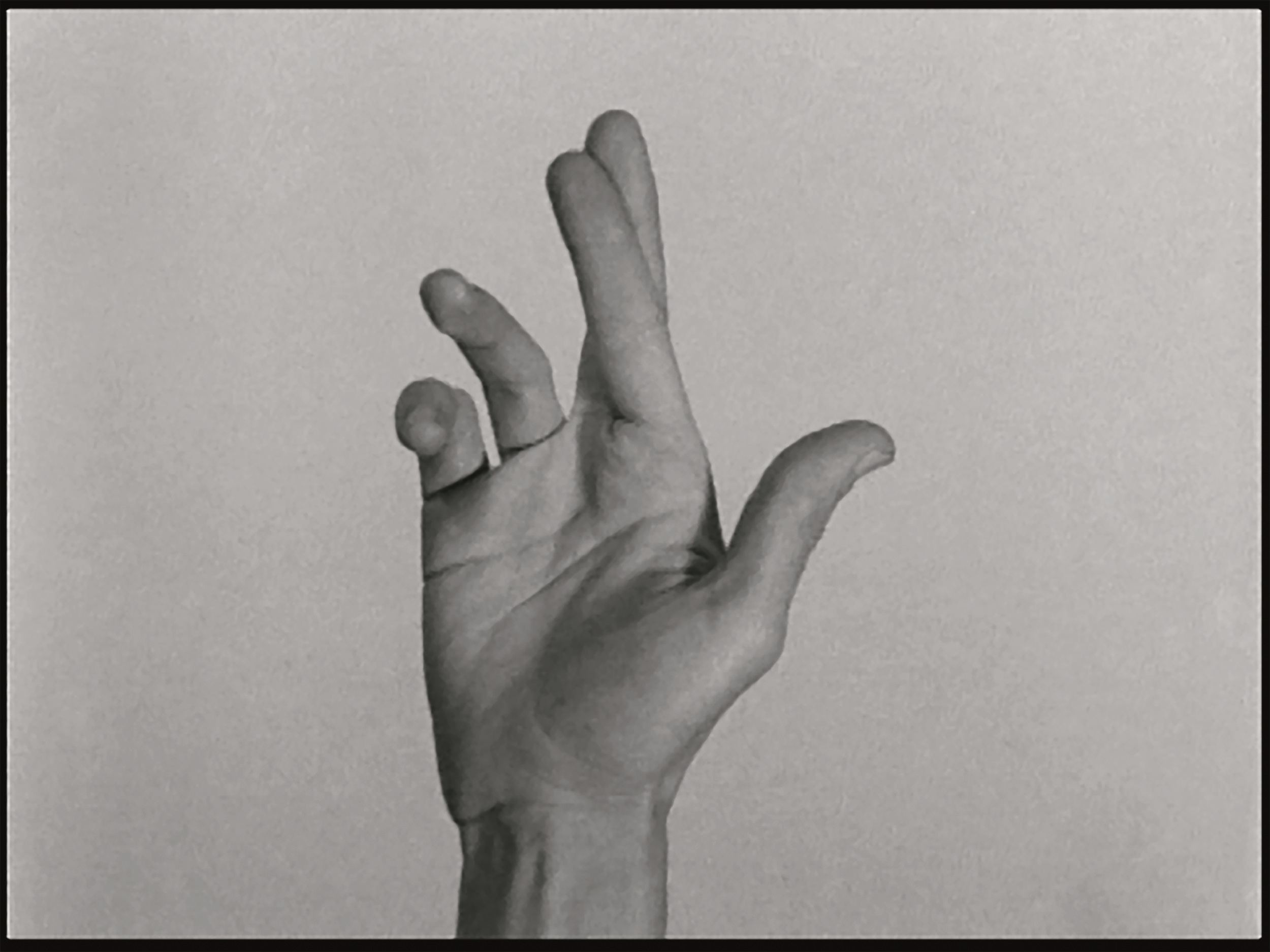 Black-and-white film still of a hand held up in the air against a blank background, palm facing forward, the first two fingers crossed.