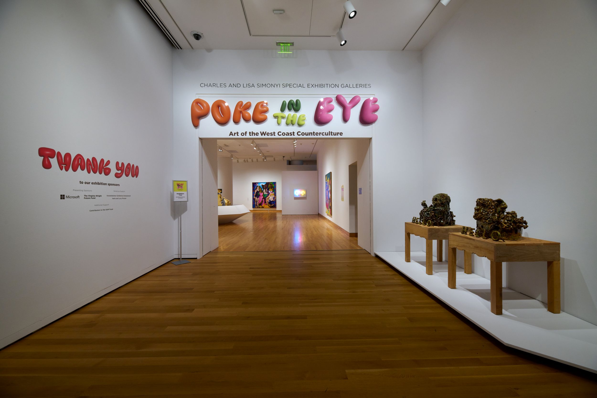 Interior of museum gallery showing entrance to an exhibition. Over the doorway, in colorful bubble letters, reads "Poke in the Eye," under which, in sans serif black text, is "Art of the West Coast Counterculture." to the right are two wooden tables with abstract sculptures; to the left bubble letters in red read "Thank you," will illegible text beneath.
