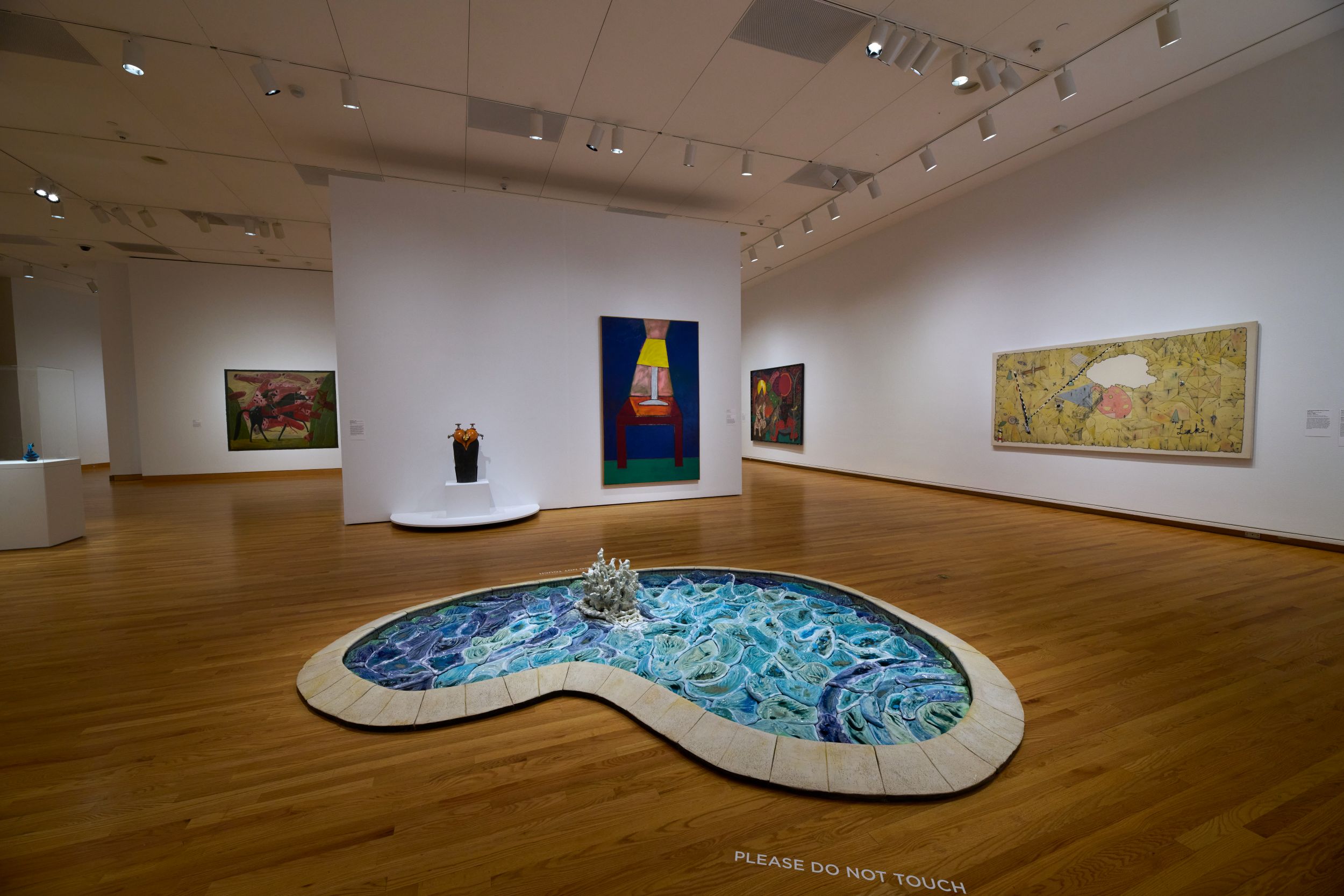 Interior of a museum gallery with white walls and wooden floors. In the center of the floor is a sculpture that mimics the form of a kidney-shaped swimming pool with a splash emerging from it; on the walls are several large abstract artworks and one small sculpture on a base.