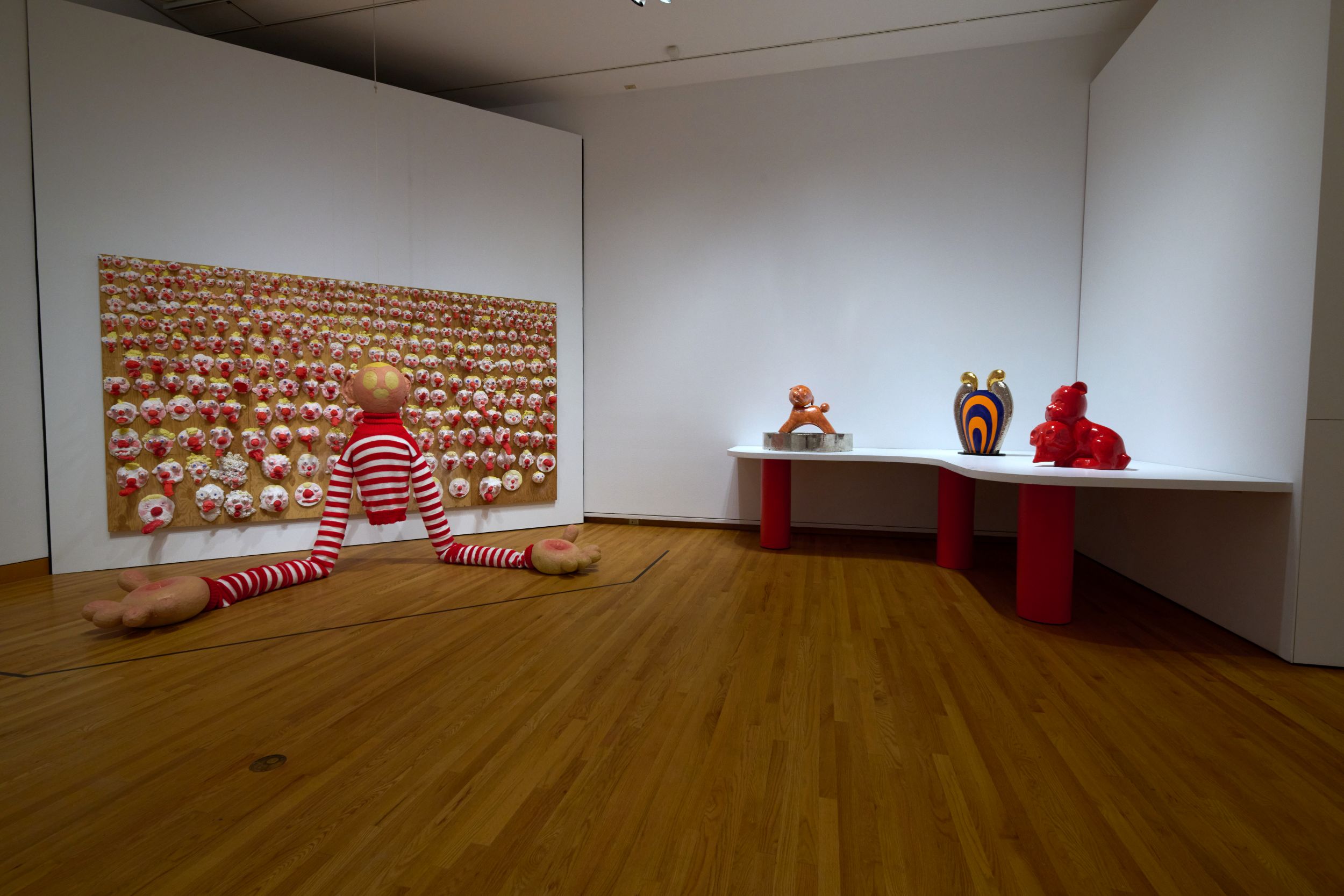 Interior of a museum gallery with white walls and wood floors. On the left wall is a rectangular artwork covered with dozens of malformed clown faces; in front of it is a torso in a striped shirt with long arms that drag on the ground, and a head with circular yellow features. To the right, three small abstract sculptures rest on a table-like base.