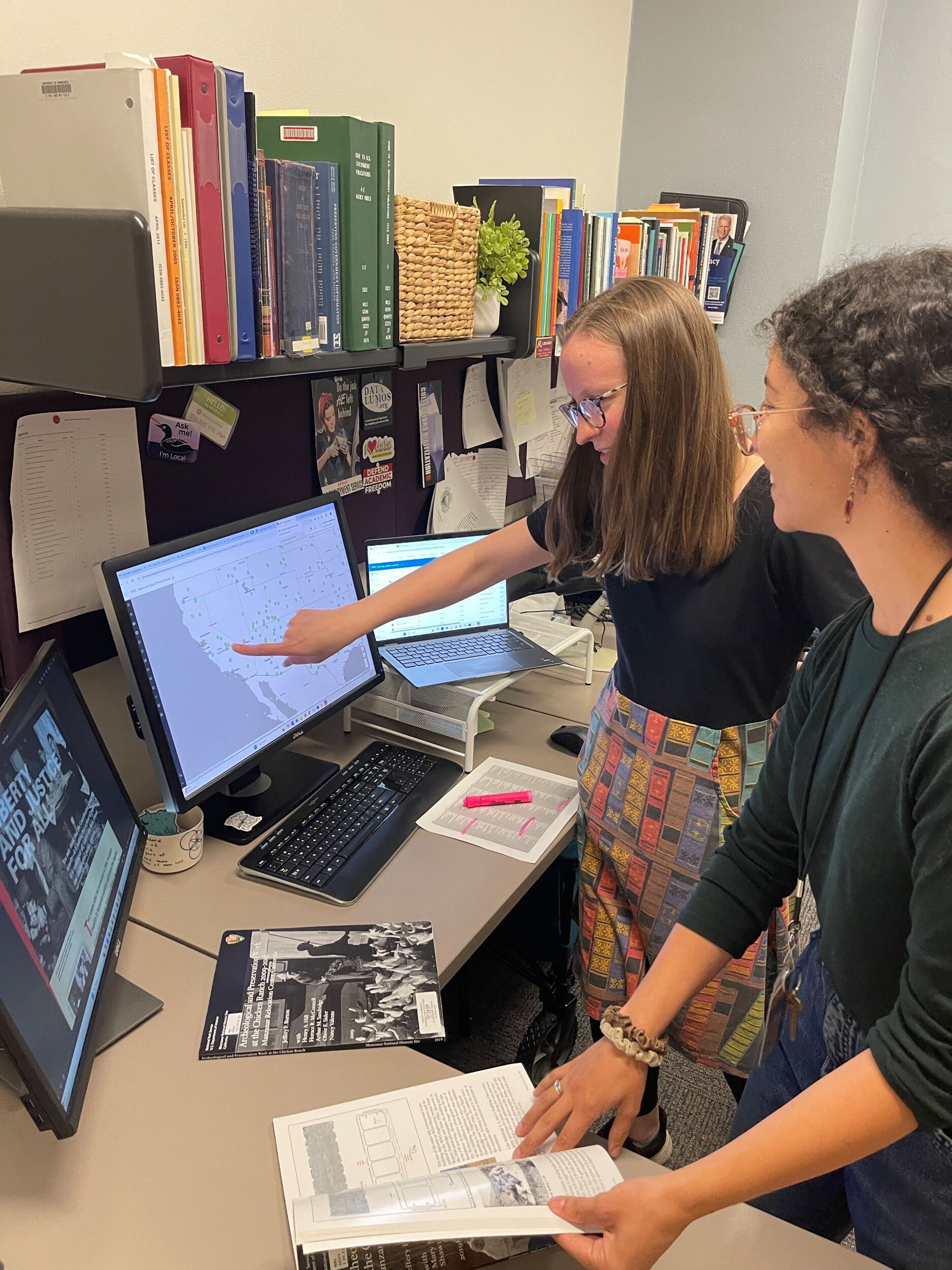Interior of an office with two women looking at a computer screen with a map of the Western United States. One woman points at the screen while the other holds a book open in front of her.