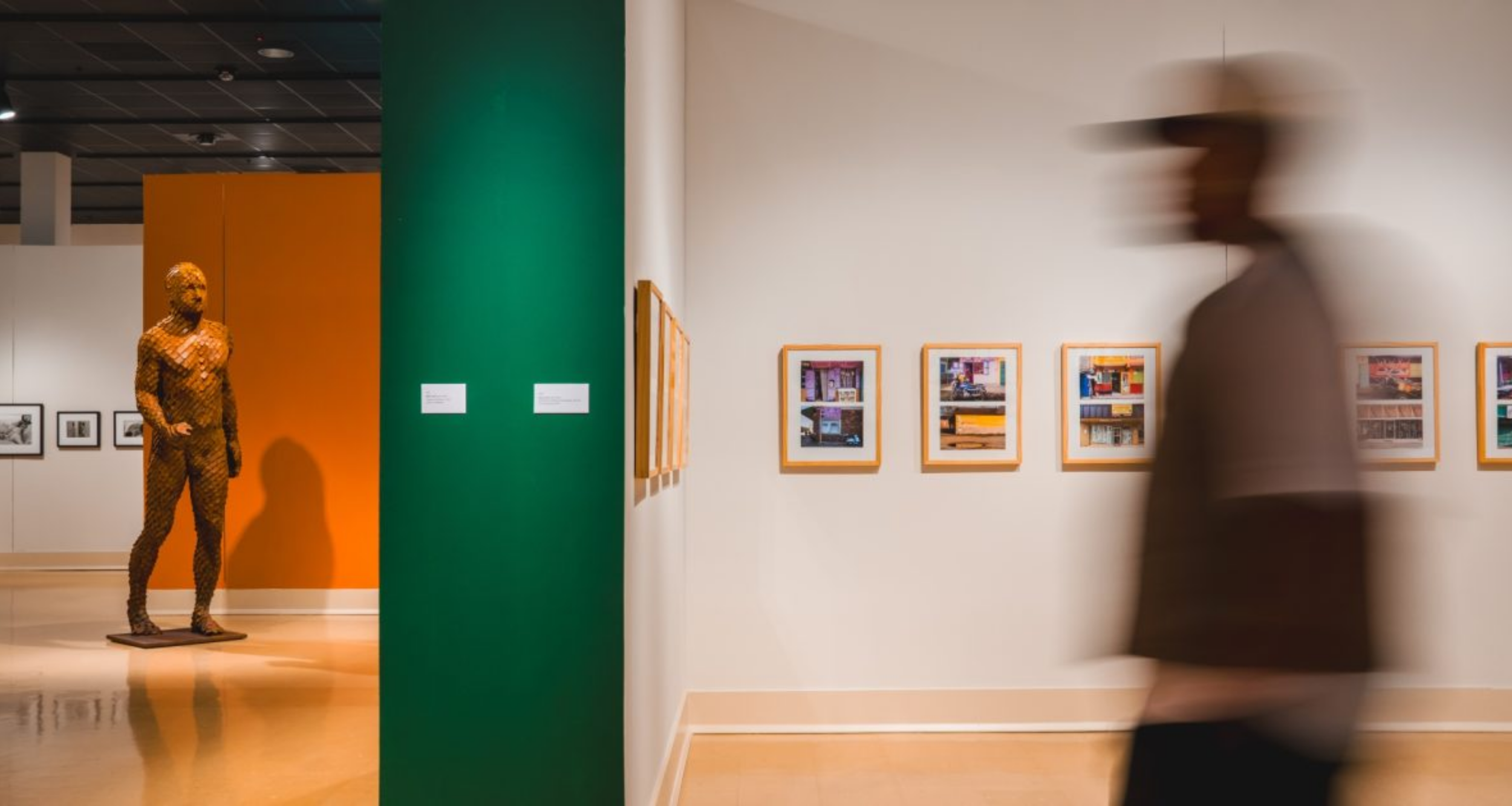 Interior of museum gallery showing a partial wall with a green endcap separating two spaces. On the left, a life-size orange sculpture of a man is posed as if to walk into the wall; on the right is a blurred long-exposure image of an actual man walking. Behind them both, framed works of art are hung on the walls.