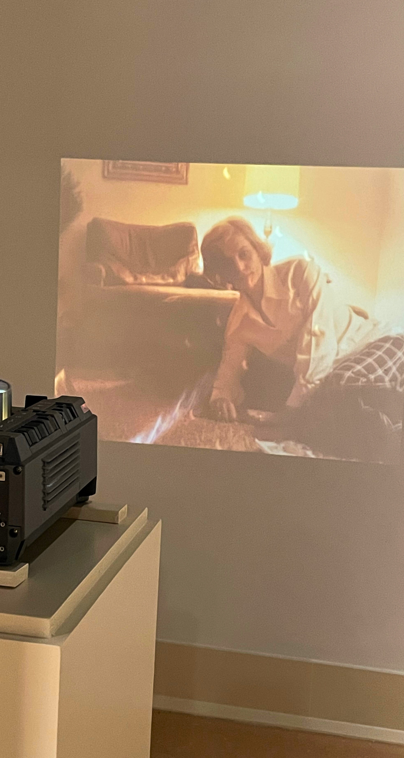 A small black film projector on a pedestal projects onto a wall an image of a person in plaid pants and a white shirt slumping on the floor in a domestic space, with an armchair and a lit lamp behind them.