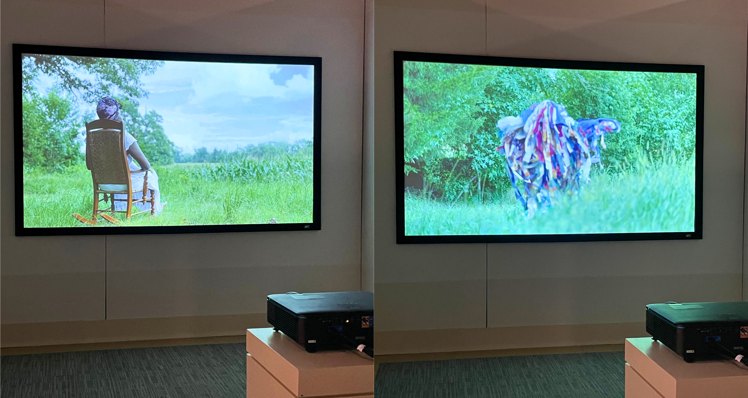 Two small black projectors on pedestals project two separate images on the opposing wall. On the left, a Black woman wearing a dress and a headwrap sits in a rocking chair in a green landscape, looking out into the distance. On the right, a person totally hidden within a costume of multicolored scraps of fabric walks through tall grass, with green bushes behind.