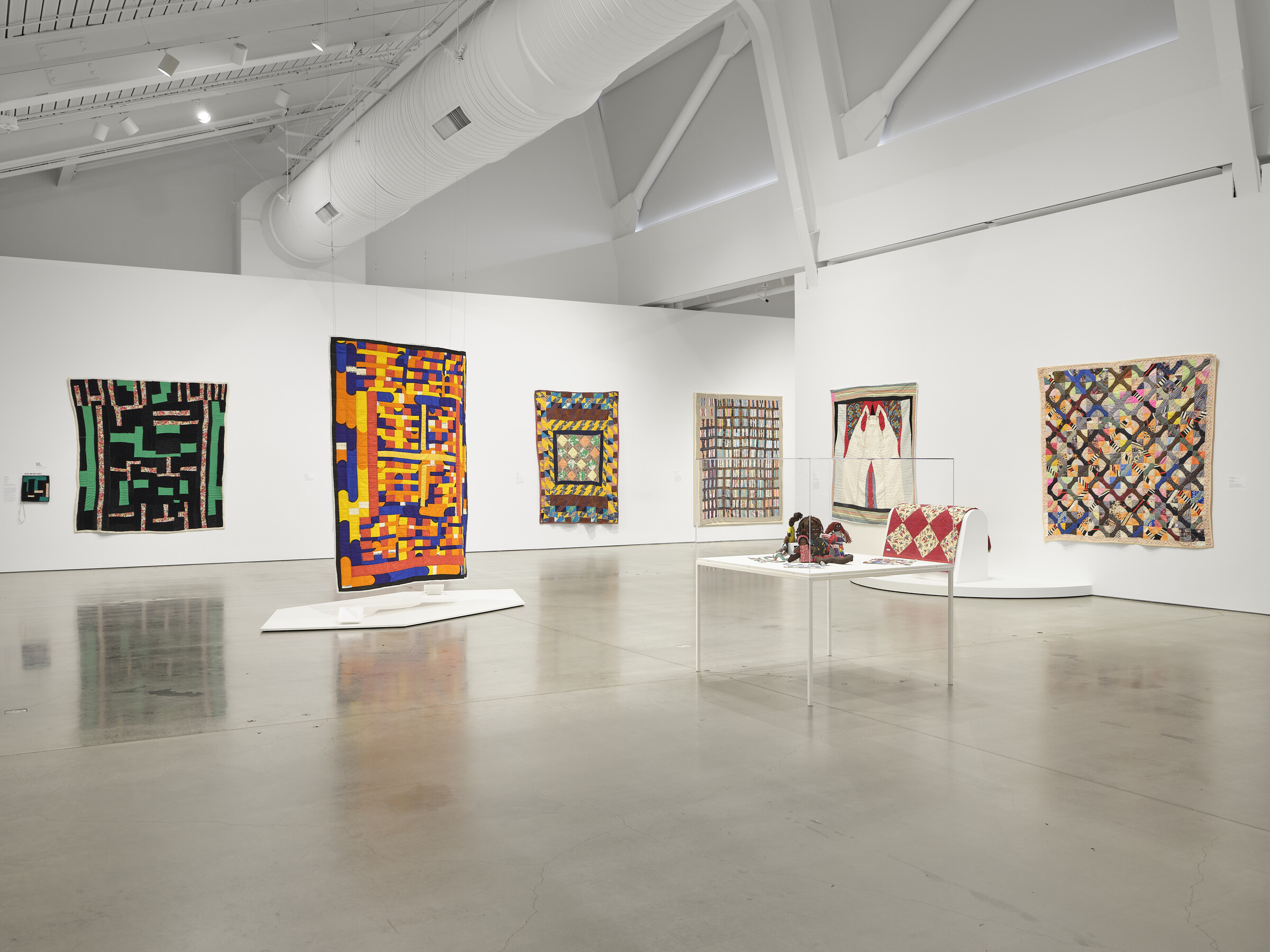 Interior of museum gallery with white walls and polished floor. Several colorful quilts are hung on the walls; one is suspended from the ceiling, and another drapes over a piece of exhibition furniture. There is also a glass case with an object in the foreground.