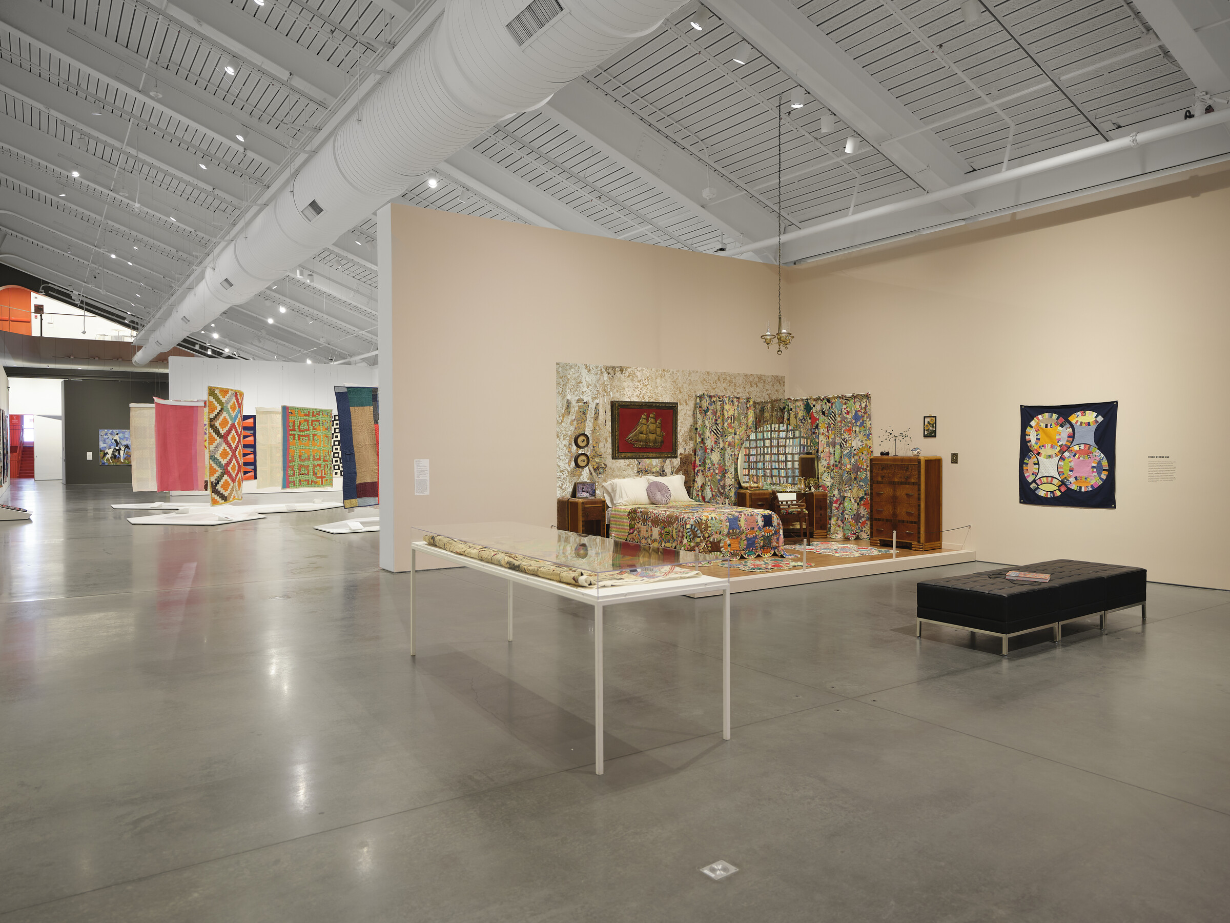 Interior of a museum gallery with an industrial ceiling and polished floors. In the foreground are two freestanding walls joined together to form a corner; they are painted a pale peach color. A bedroom is set up in the corner, with a bureau, bed, nightstand, and dressing table, as well as many quilts.