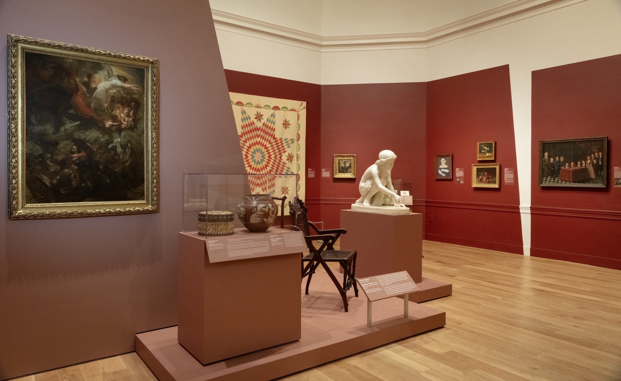 Interior of museum gallery with red-painted walls. Objects on view include paintings, a quilt, ceramic vessels, a chair, and a marble statue.