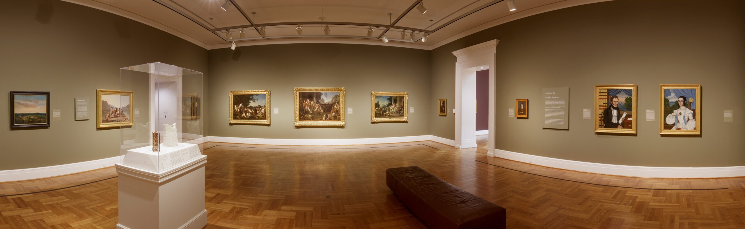 Panoramic view of a museum gallery with polished parquet floors and sage-green painted walls hung with paintings.